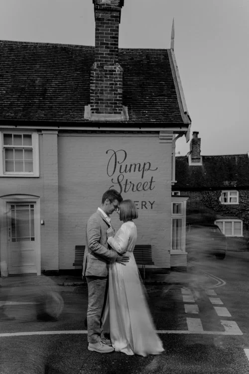 A couple share a quiet, intimate embrace outside the quaint Pump Street Bakery in a charming village setting, captured in evocative black and white. Their gentle moment contrasts beautifully with the soft blur of passers-by moving around them, creati