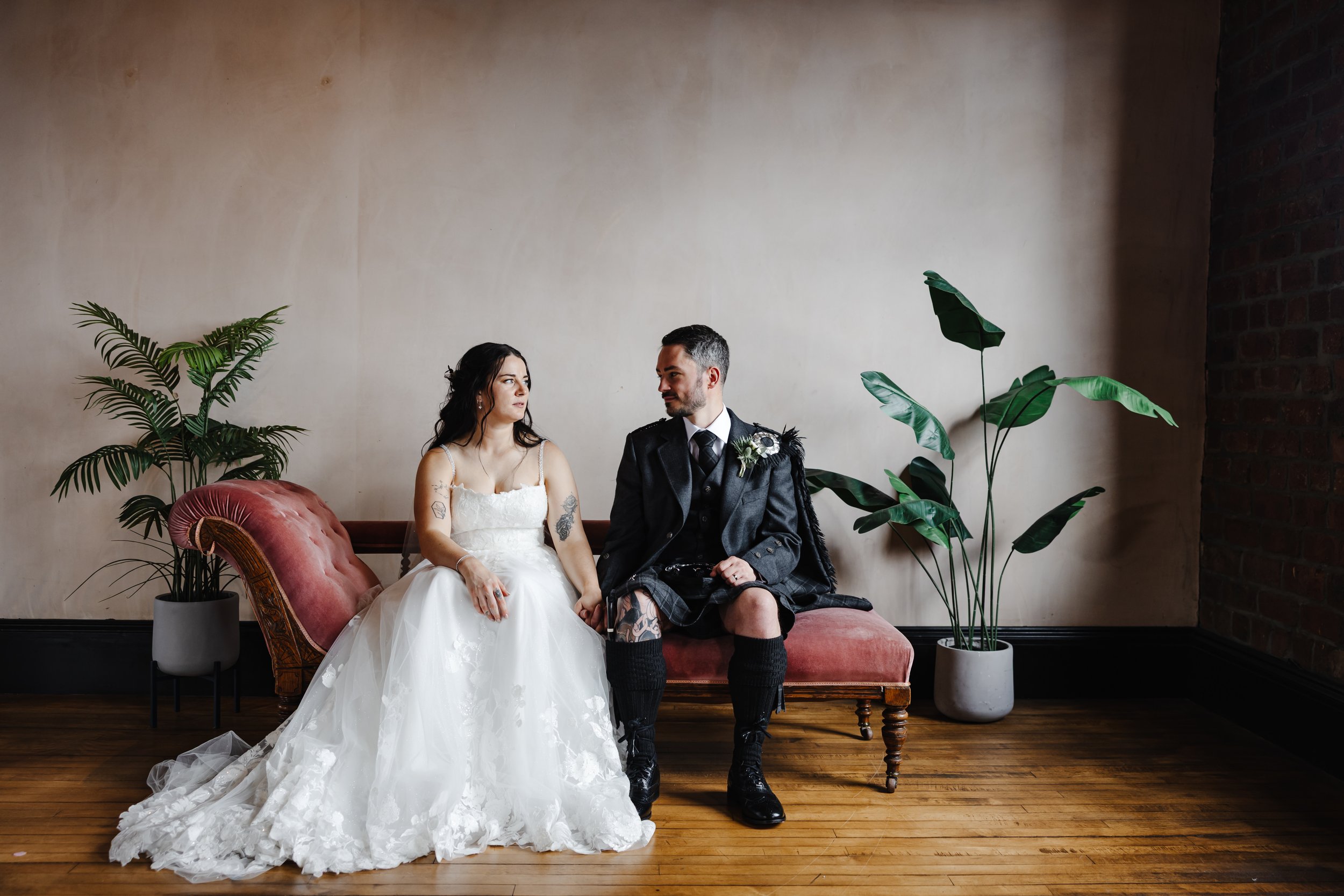 Marie and Oliver sat on a red chaise longue. Marie wears a white wedding dress, while Oliver wears a shirt, tie, blazer, and kilt.