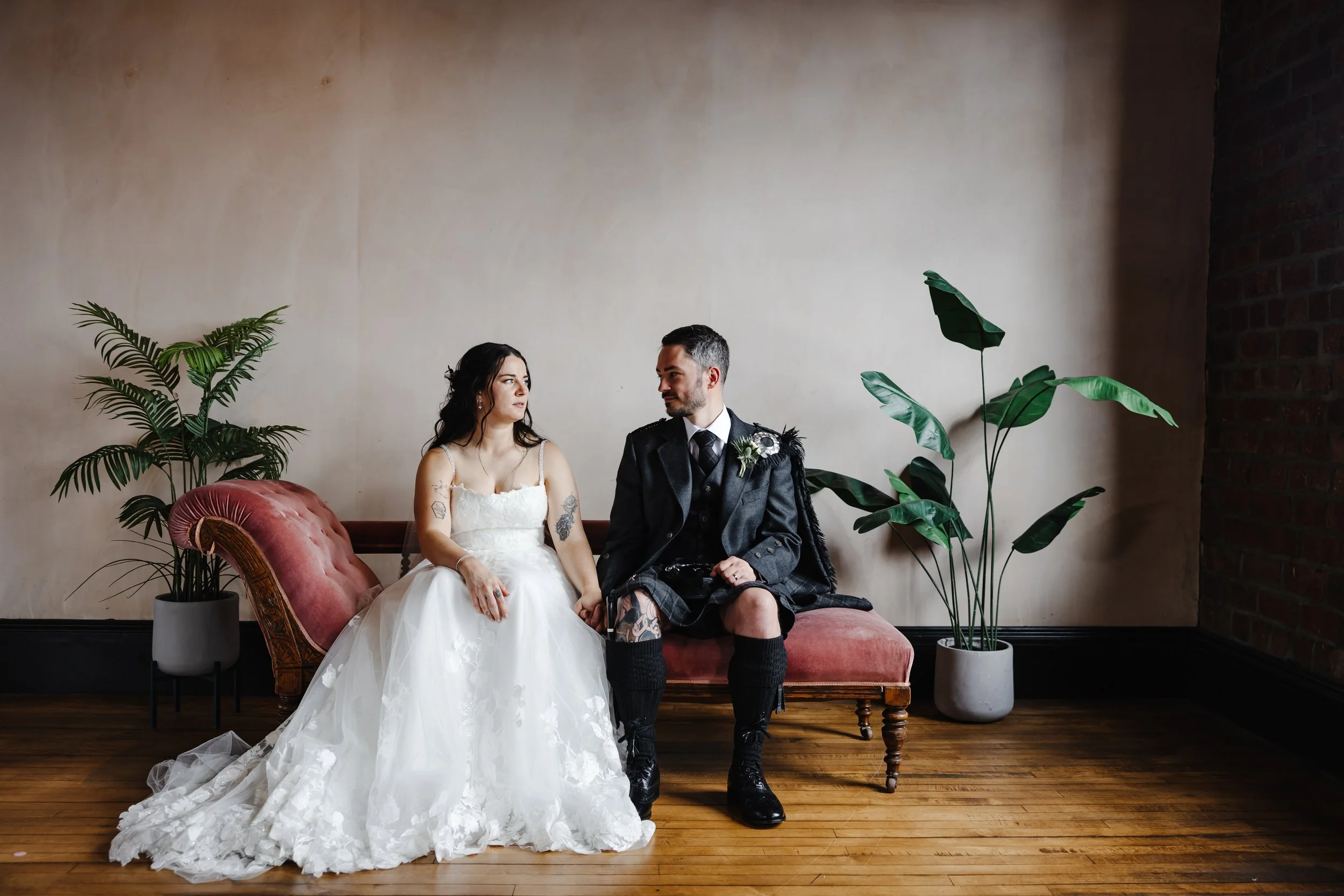 Marie and Oliver sat on a red chaise longue. Marie wears a white wedding dress, while Oliver wears a shirt, tie, blazer, and kilt.
