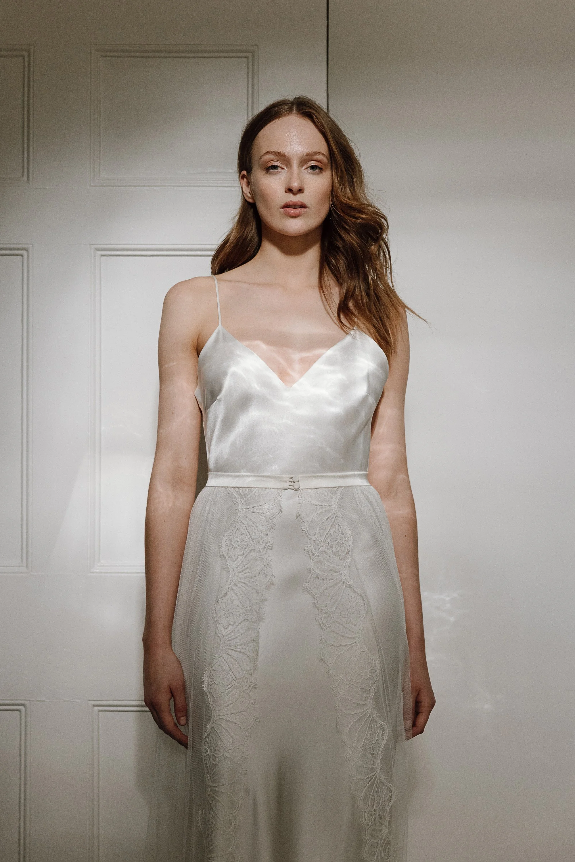 Soft light falls across the bride as she stands against a paneled white wall, wearing a fluid silk slip dress with the faintest sheen, its fine spaghetti straps framing a gentle V neckline that flows into a skirt detailed with delicate lace panels th