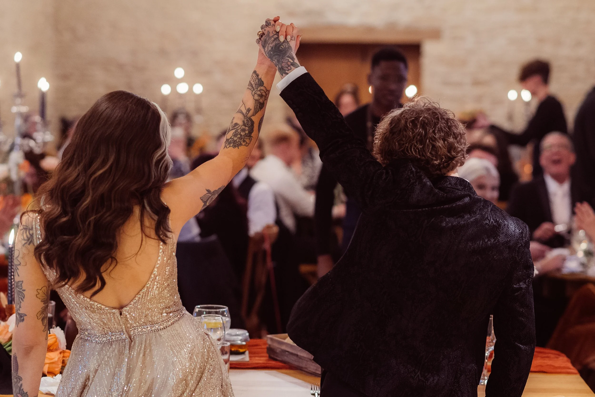 A joyful wedding reception moment captured by Gemma Gaskins Photography as the newlyweds raise their joined hands to celebrate with their guests. The bride’s shimmering gown, their matching tattoos and the warm candlelit atmosphere create a stylish a
