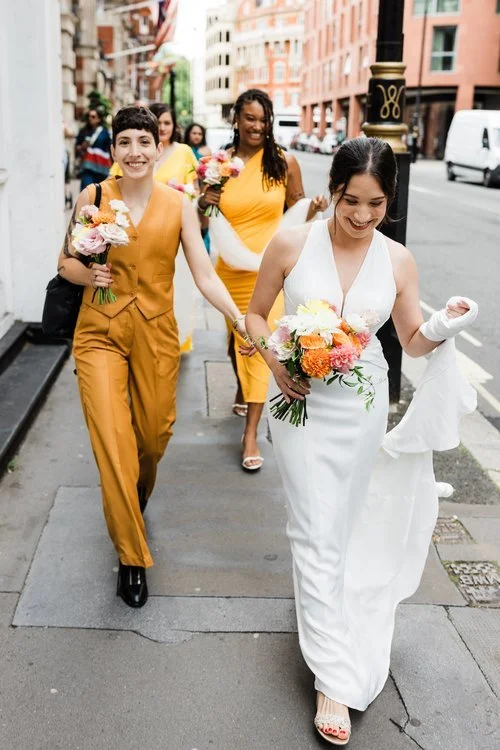 Modern city wedding party carrying vibrant seasonal bouquets by Botanique Workshop featuring colourful dahlias, roses and fresh garden foliage. With the bride and bridal party walking through London streets in stylish yellow and white outfits, this j