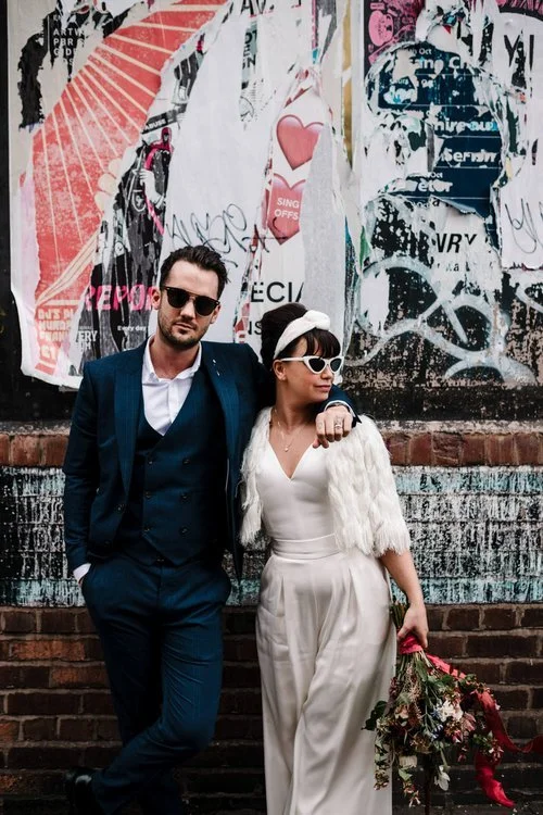 A bold and contemporary wedding portrait captured by Amy Faith Photography, showing the couple striking a confident pose against a wall of layered street posters and edgy urban textures. The groom’s tailored navy suit and relaxed stance create a cool