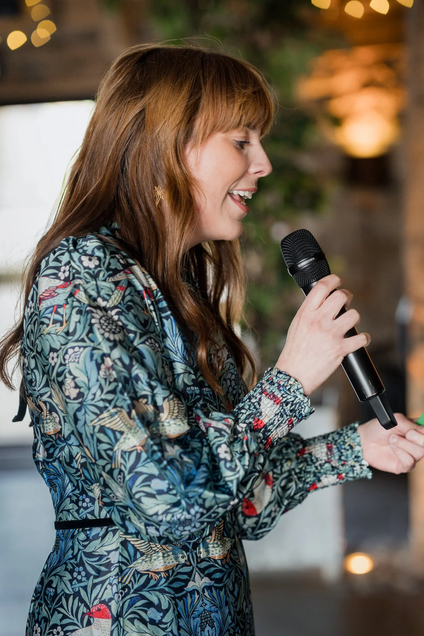 Abi, the Full Of Soul Celebrant is holding a microphone during a wedding ceremony and wearing a green, paisley dress.