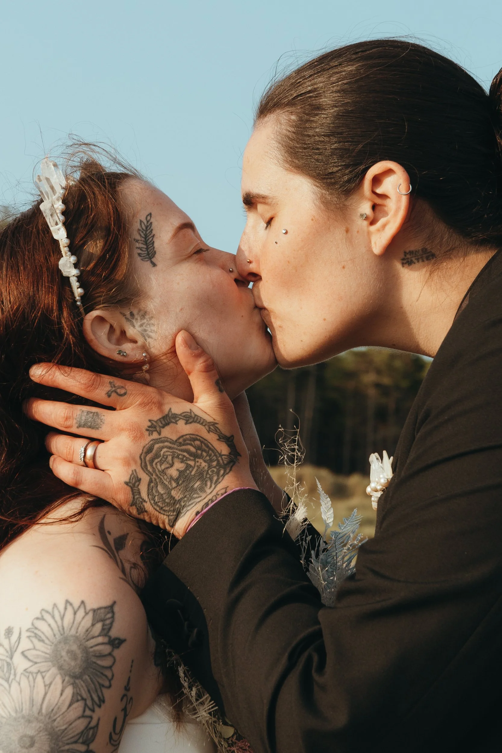 Two people sharing a tender kiss during their wedding ceremony. They both have visible tattoos and piercings, adding a personal and expressive touch to the image. One person gently holds the other’s face, their wedding rings and hand tattoos clearly 