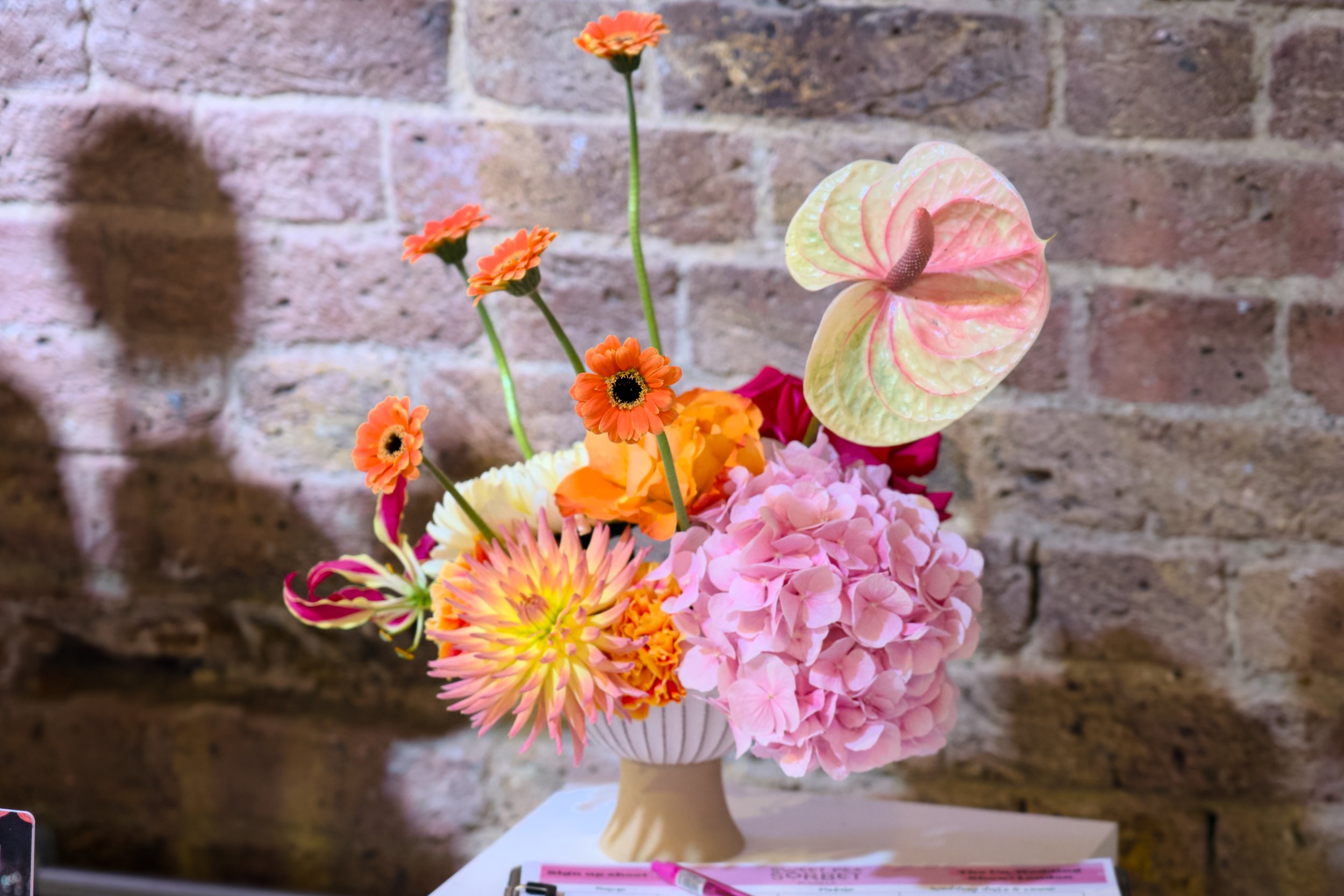 Vibrant floral arrangement by Sweet Pea Sorbet Floristry featuring orange gerberas, pink hydrangeas, peach dahlias, and a blush anthurium in a sculptural vase against a rustic brick wall. A bold and modern design perfect for weddings or events.