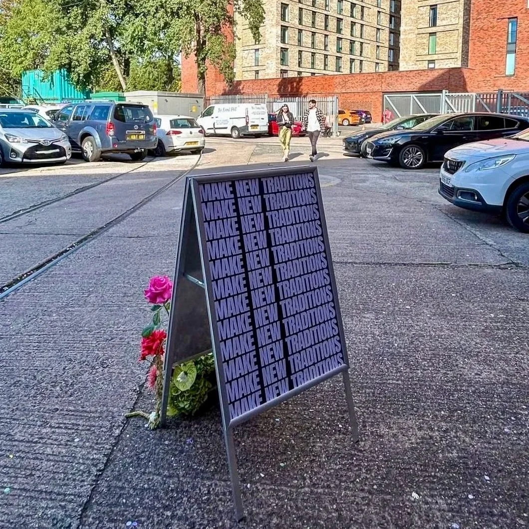 A sign outside a wedding fair that says MAKE NEW TRADITIONS repeatedly on it. There are some roses in front of it.