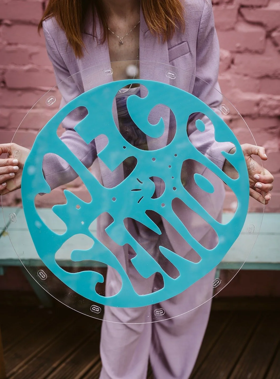 Wedding celebrant Meg Senior in a lilac suit holds a circular acrylic sign with bold turquoise lettering spelling ‘Meg Senior.’ The modern, playful sign stands out against a pink brick wall, showcasing a stylish and creative wedding brand aesthetic. 