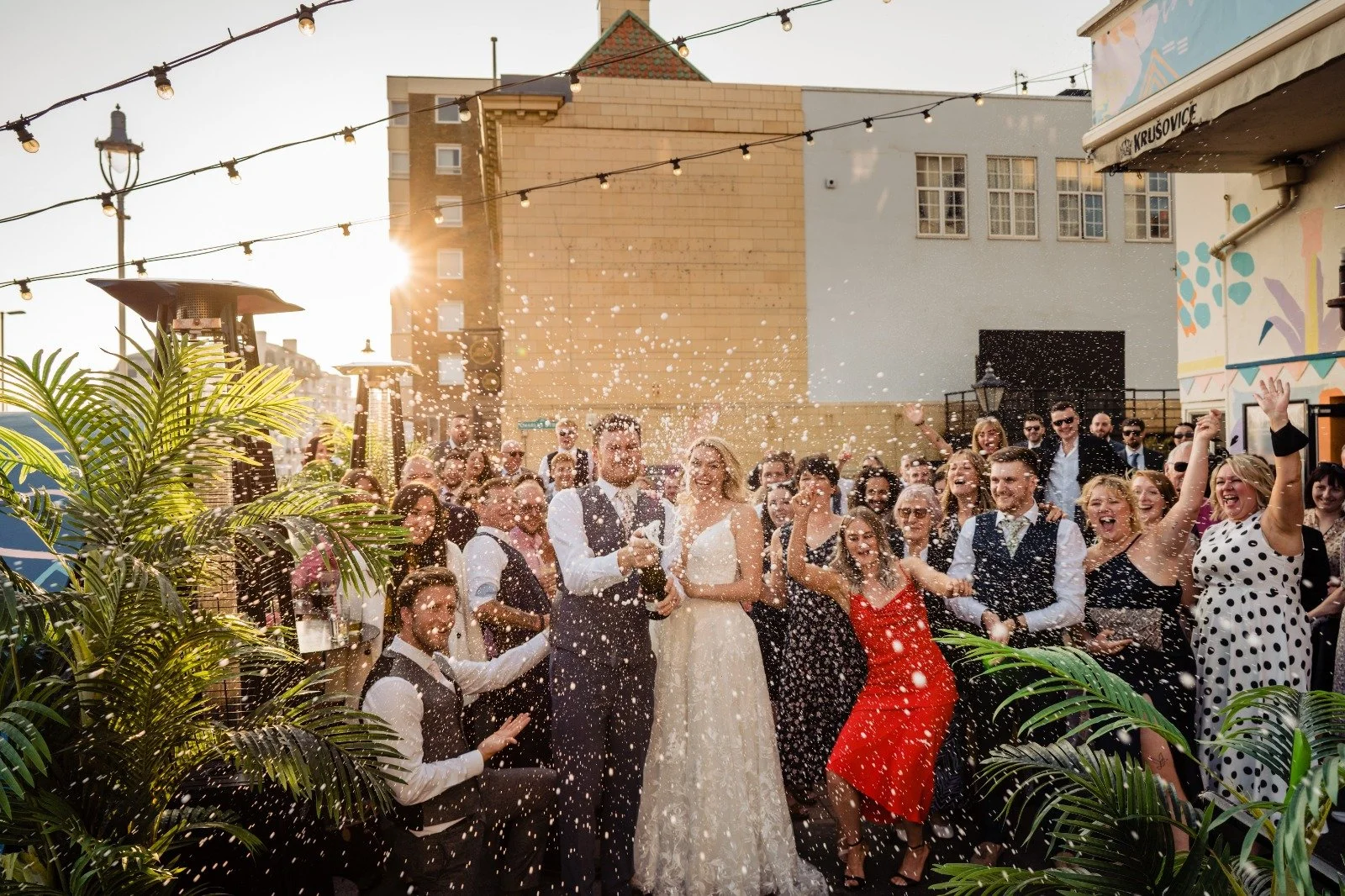 Bride and groom celebrate with guests outdoors at Pier Court Wedding Venue in Brighton, capturing a joyful, sunlit seaside wedding moment beneath festoon lights.