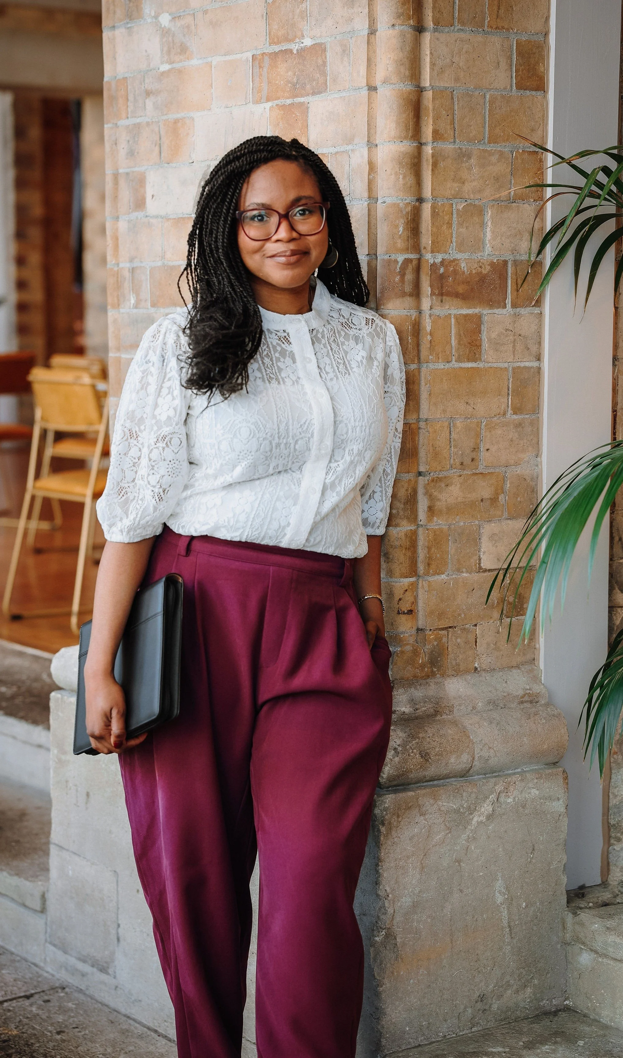 A Black wedding celebrant called Deyi, founder of Gather & Tell Celebrations is wearing a pretty blouse and red trousers. She's smiling at the camera just before a ceremony. Photo by Elly Deakin.
