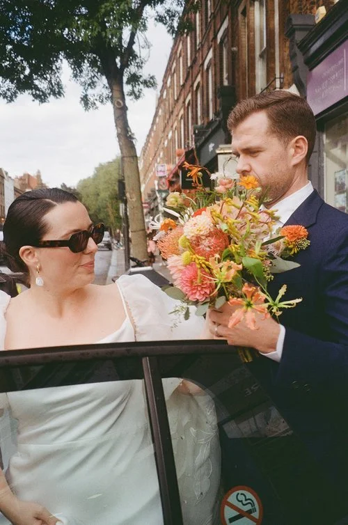 A bride steps out of a car onto a bustling London street as her partner waits beside her, holding a vibrant bouquet of dahlias, roses, and textured summer blooms. Her chic sunglasses and elegant gown create a stylish contrast with the urban backdrop 