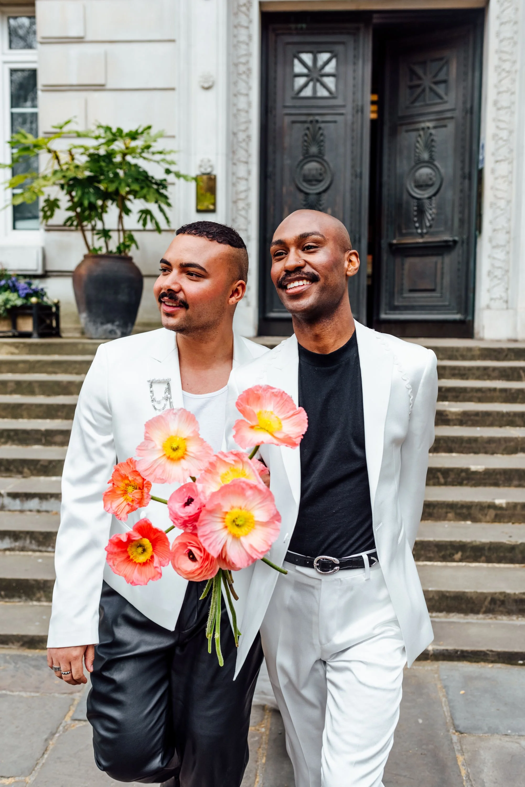 A stylish wedding portrait featuring two grooms in coordinated modern suits, captured with a vibrant bouquet of coral and yellow flowers. The fresh, contemporary look showcases Powder Blue Artistry’s natural and polished makeup style, perfect for fas