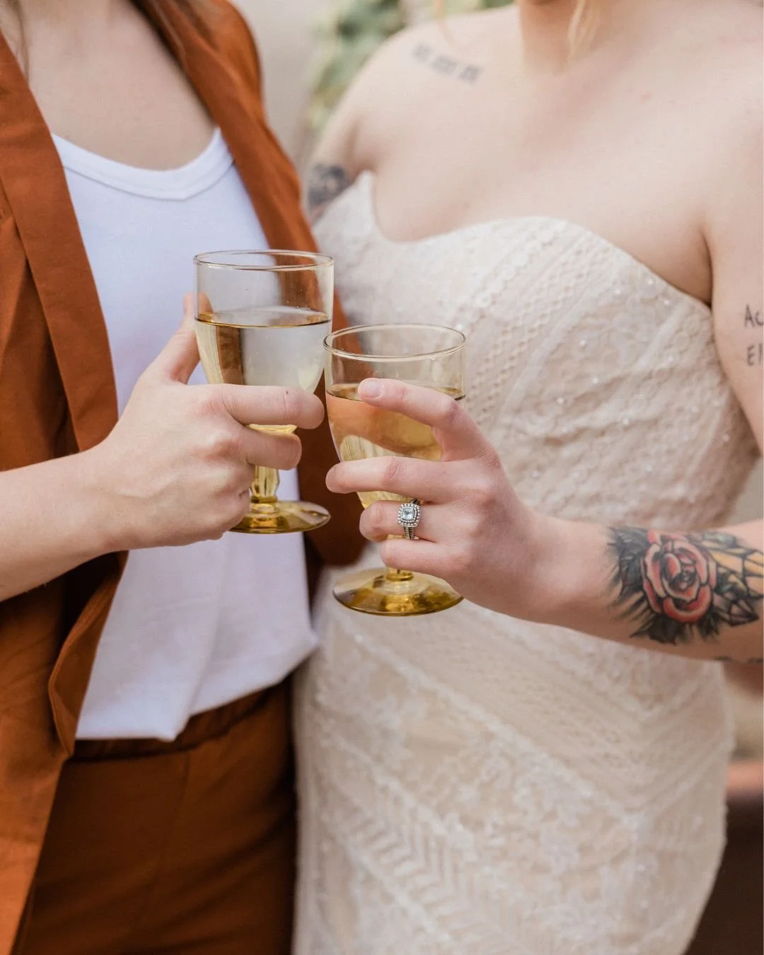A close up of a lesbian couple on their wedding day. One is wearing a beaded wedding dress and the other has a rust coloured suit. They are both holding a glass of champagne.