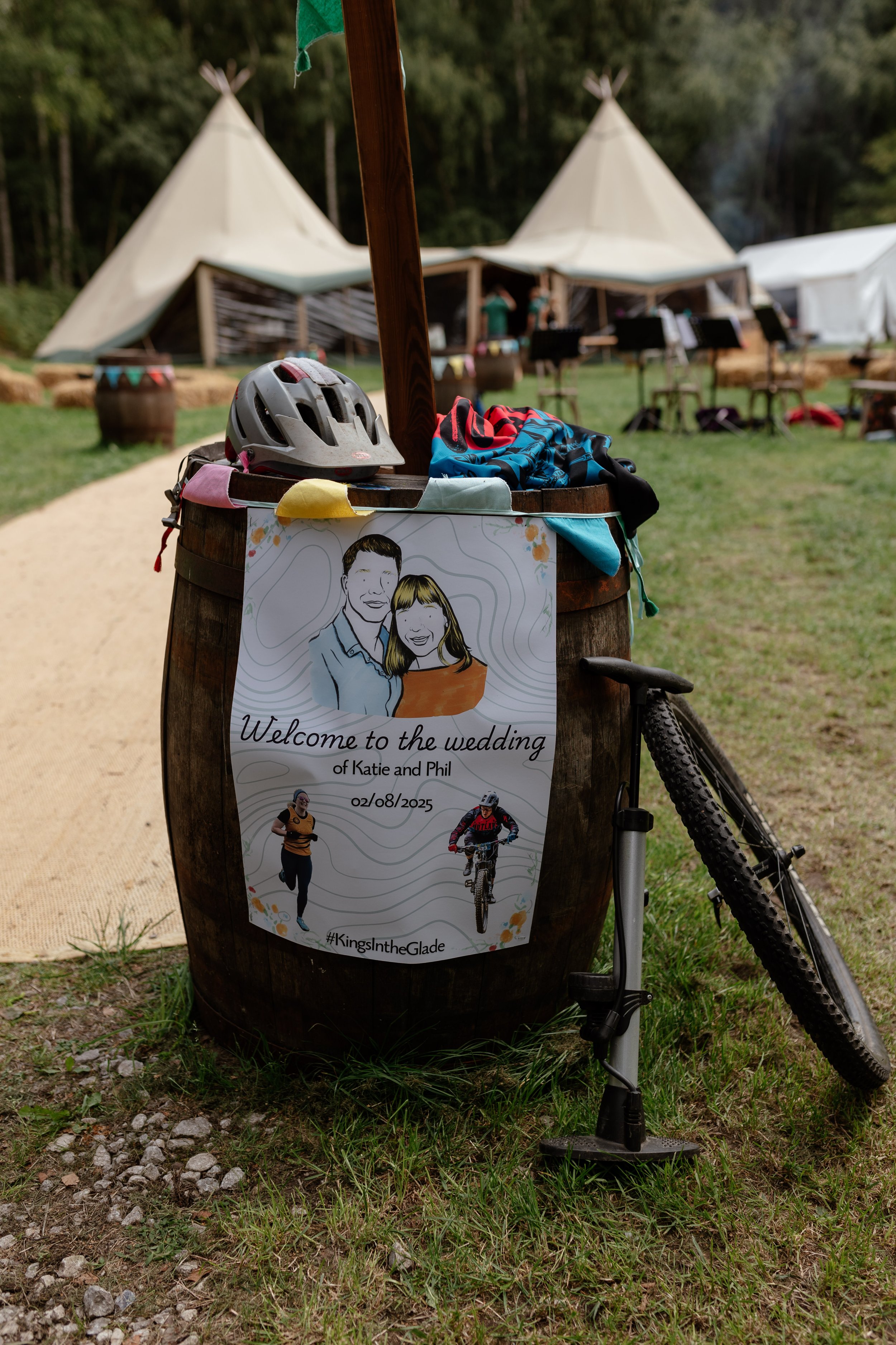 A wooden barrel with a poster that says 'welcome to the wedding of Katie and Phil' there are some cycling object placed around it.