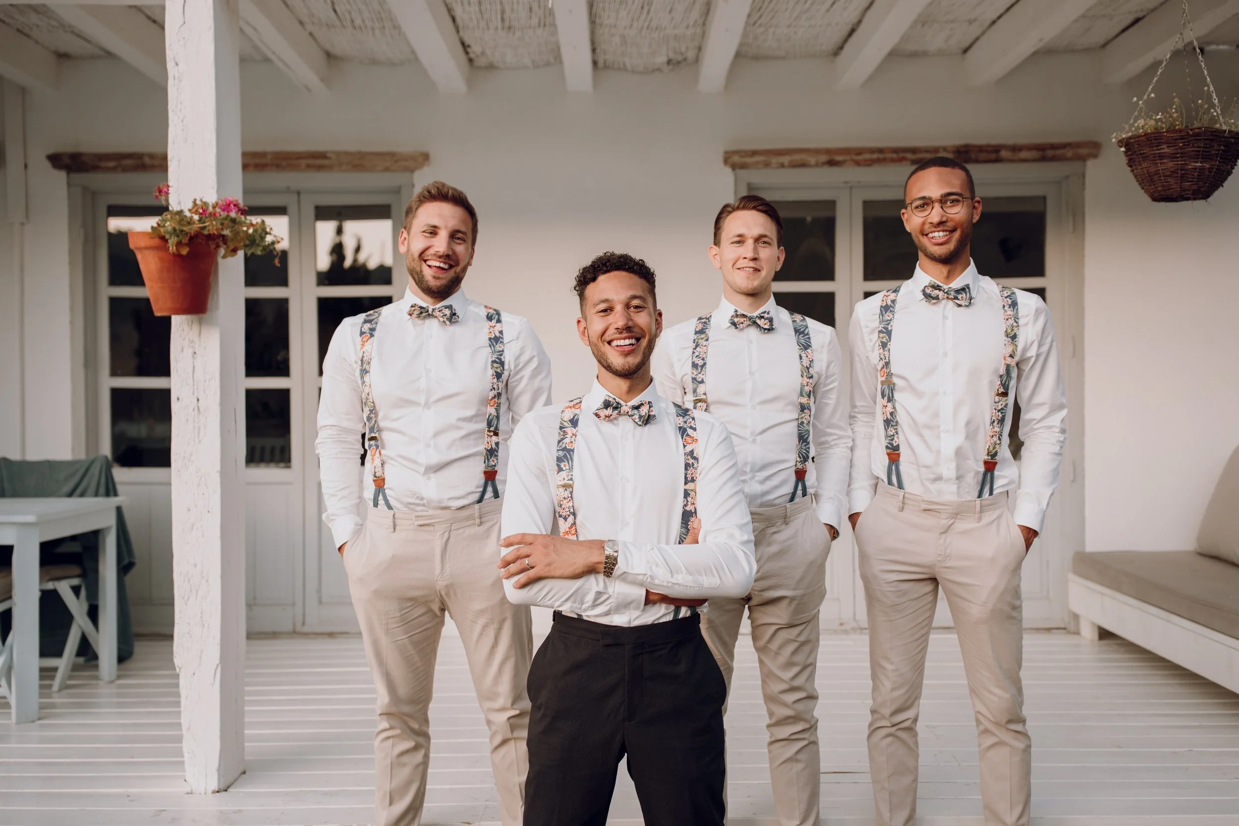 Four groomsmen standing on a white wooden terrace in front of a light coloured building, all dressed in white shirts with patterned floral suspenders and matching bow ties. Three of the men wear light beige trousers while the man in front wears darke