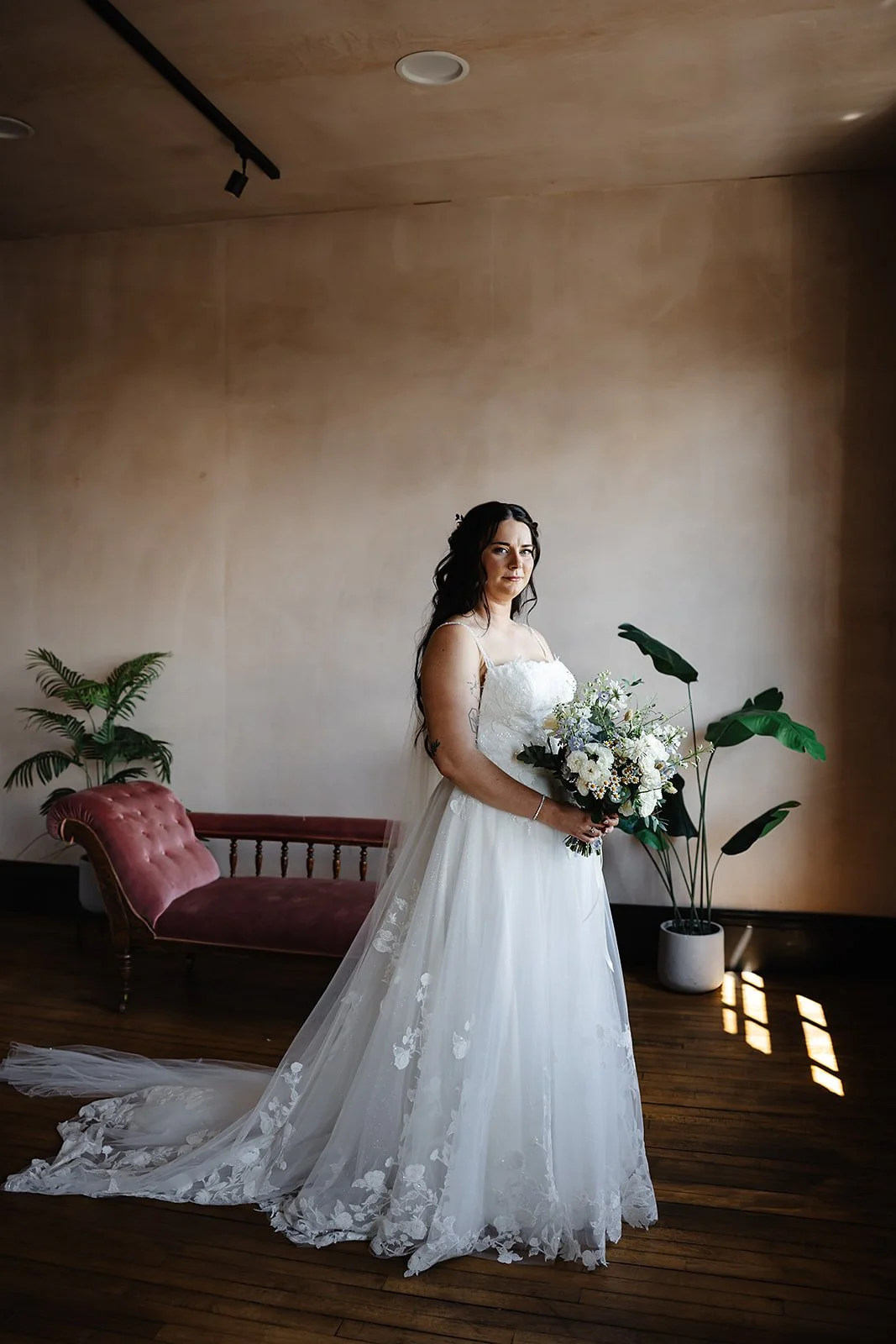 Marie in her Aimee Bridal Couture wedding dress, holding a bouquet in front of a red Chaise Longue.
