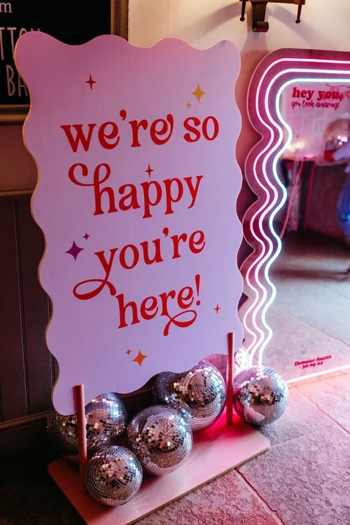 Playful wedding welcome sign by AMW Events reading “we’re so happy you’re here!” in red retro-style font, surrounded by sparkling disco balls, next to a neon-lit wavy mirror in a vibrant pink setting.









