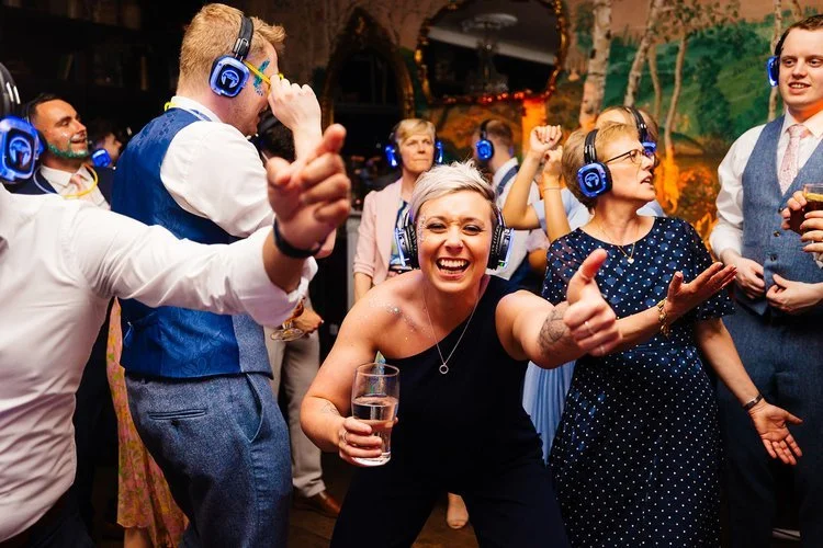 A lively silent-disco wedding party on the dance floor, with guests wearing glowing blue headphones and dancing in a decorated reception venue. A woman in the foreground smiles directly at the camera giving two thumbs up, surrounded by energetic danc