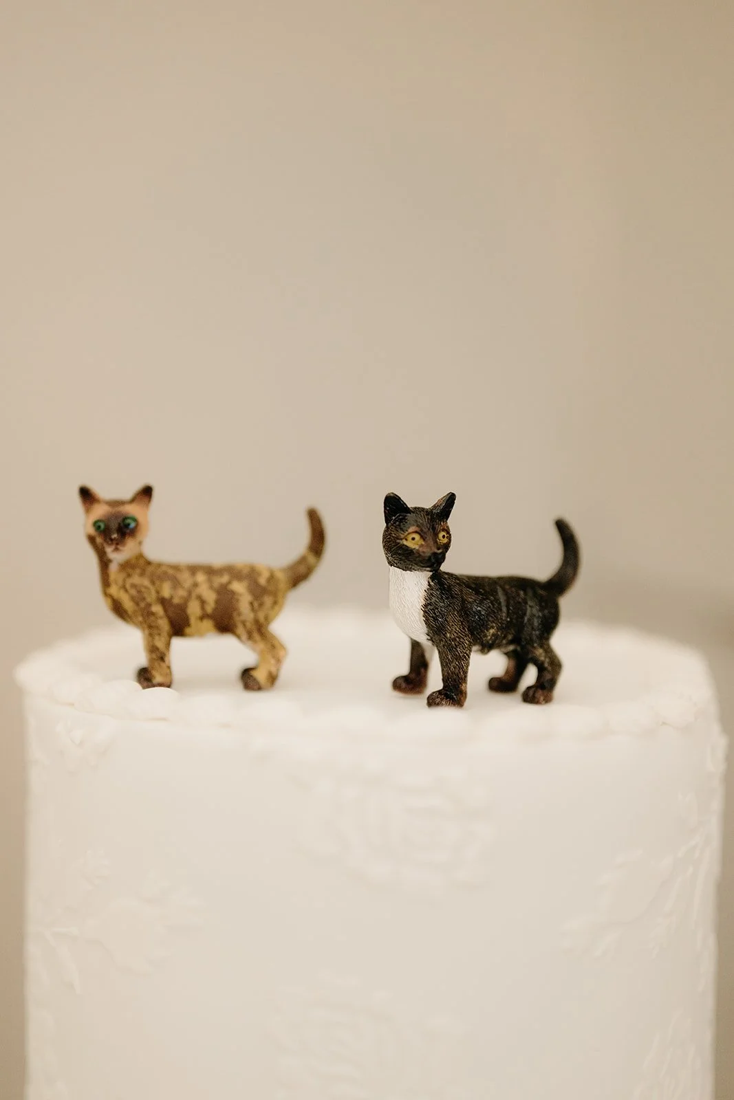 A close up of 2 cat ornaments on a white, wedding cake. One of the cats is is tortoiseshell and the other is black and white.