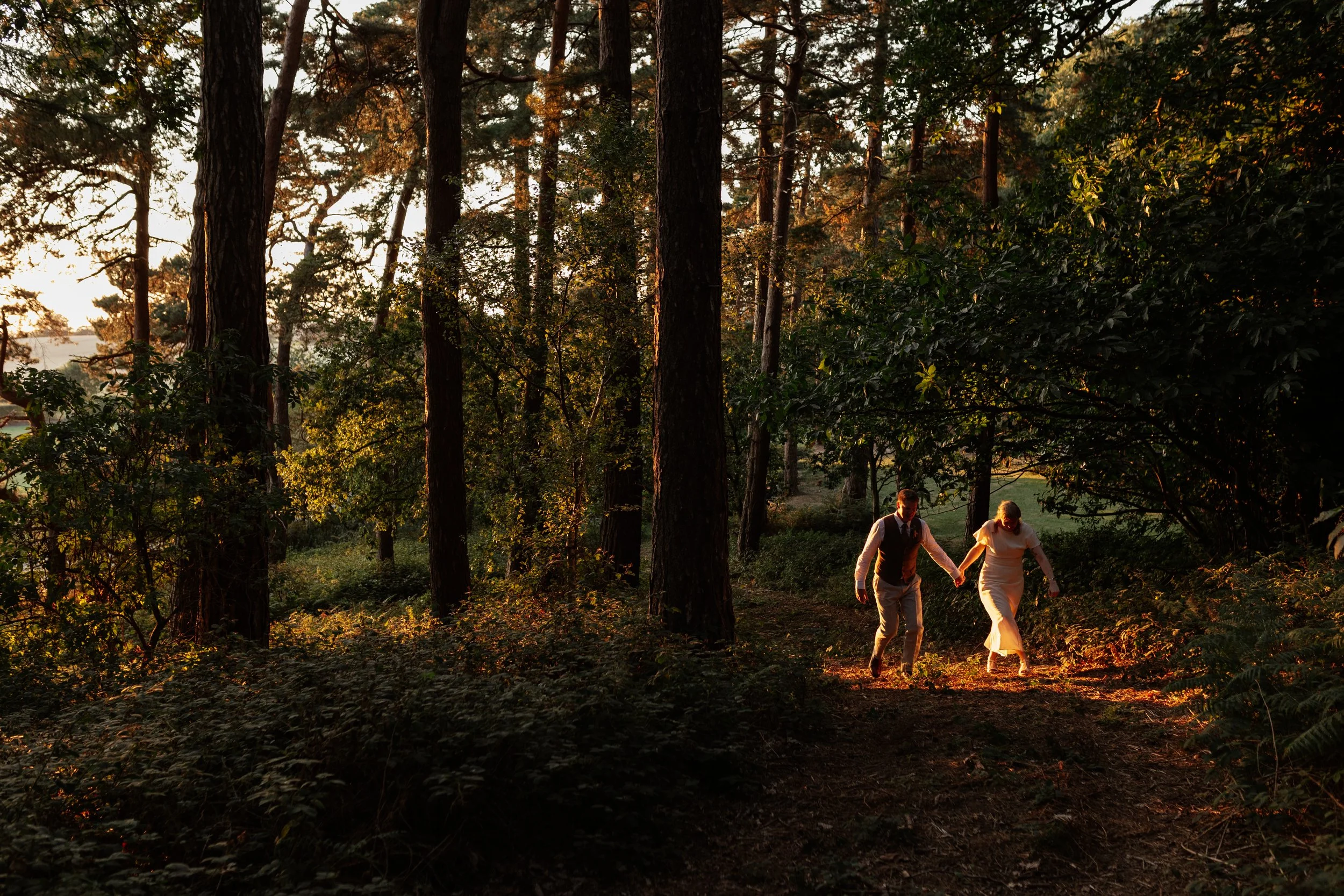 A romantic woodland wedding portrait captured by Laura Wilson Photography, showing a couple walking hand-in-hand through a sunlit forest at golden hour. The warm evening light filters through the tall trees, casting a soft glow on the couple as they 