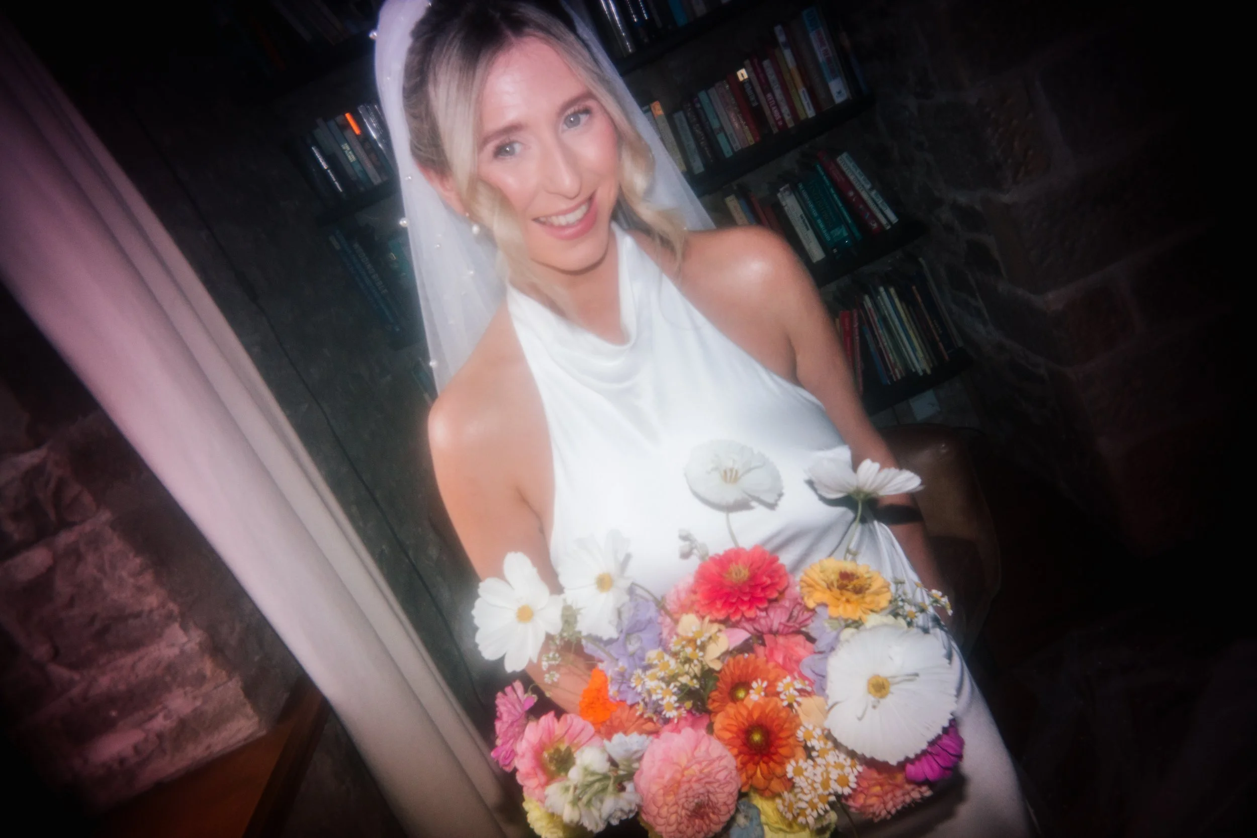 A bride in a white halter-neck wedding dress smiles warmly while holding a vibrant, colourful bouquet of summer flowers. Her veil is gently draped behind her as she stands against a backdrop of bookshelves and stone walls, creating a cosy, intimate w