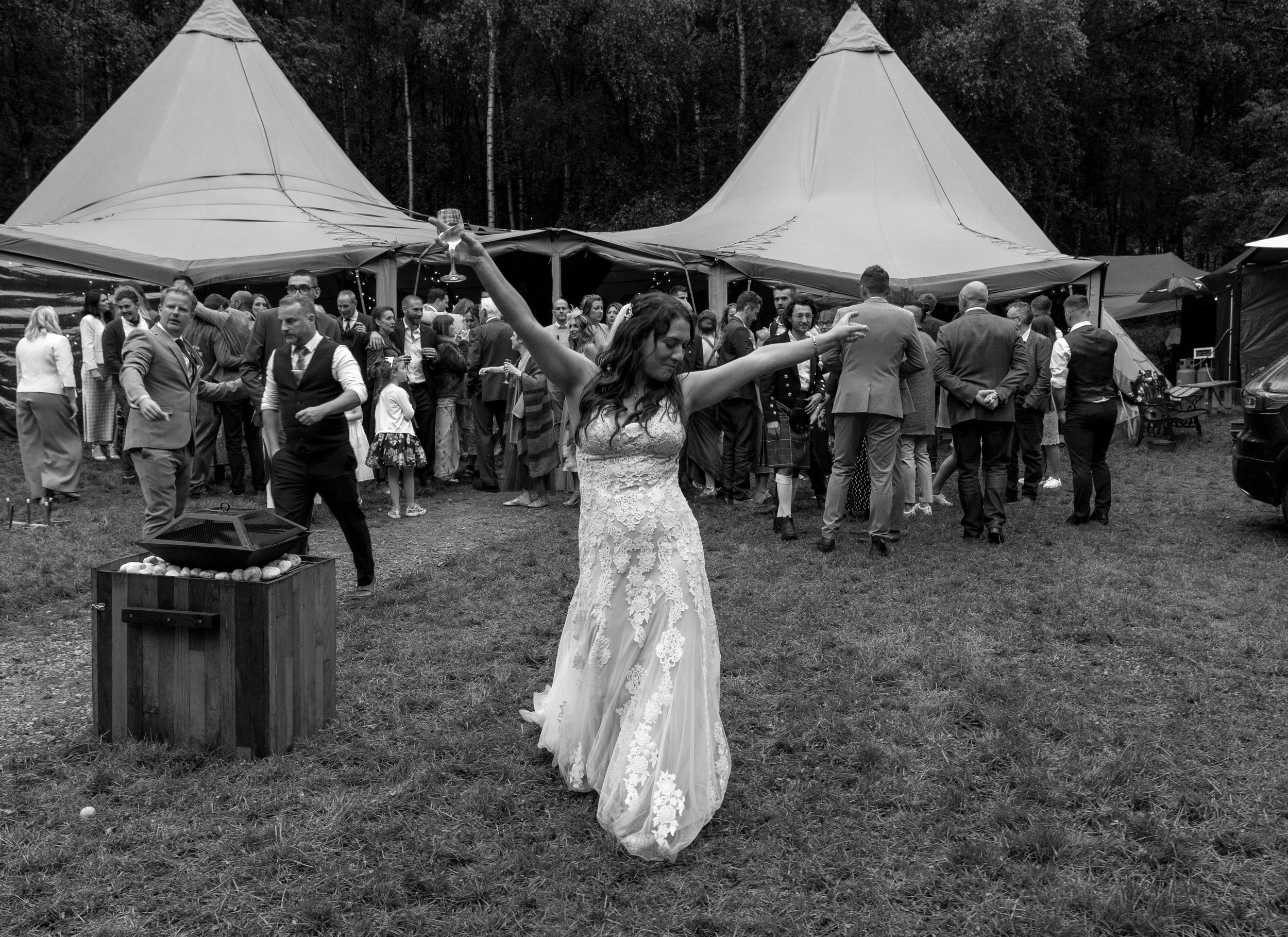 A lively black-and-white wedding moment captured by Laura Wilson Photography, showing the bride dancing with her arms outstretched in front of a bustling outdoor tipi reception. Guests gather behind her, chatting, laughing, and celebrating under the 