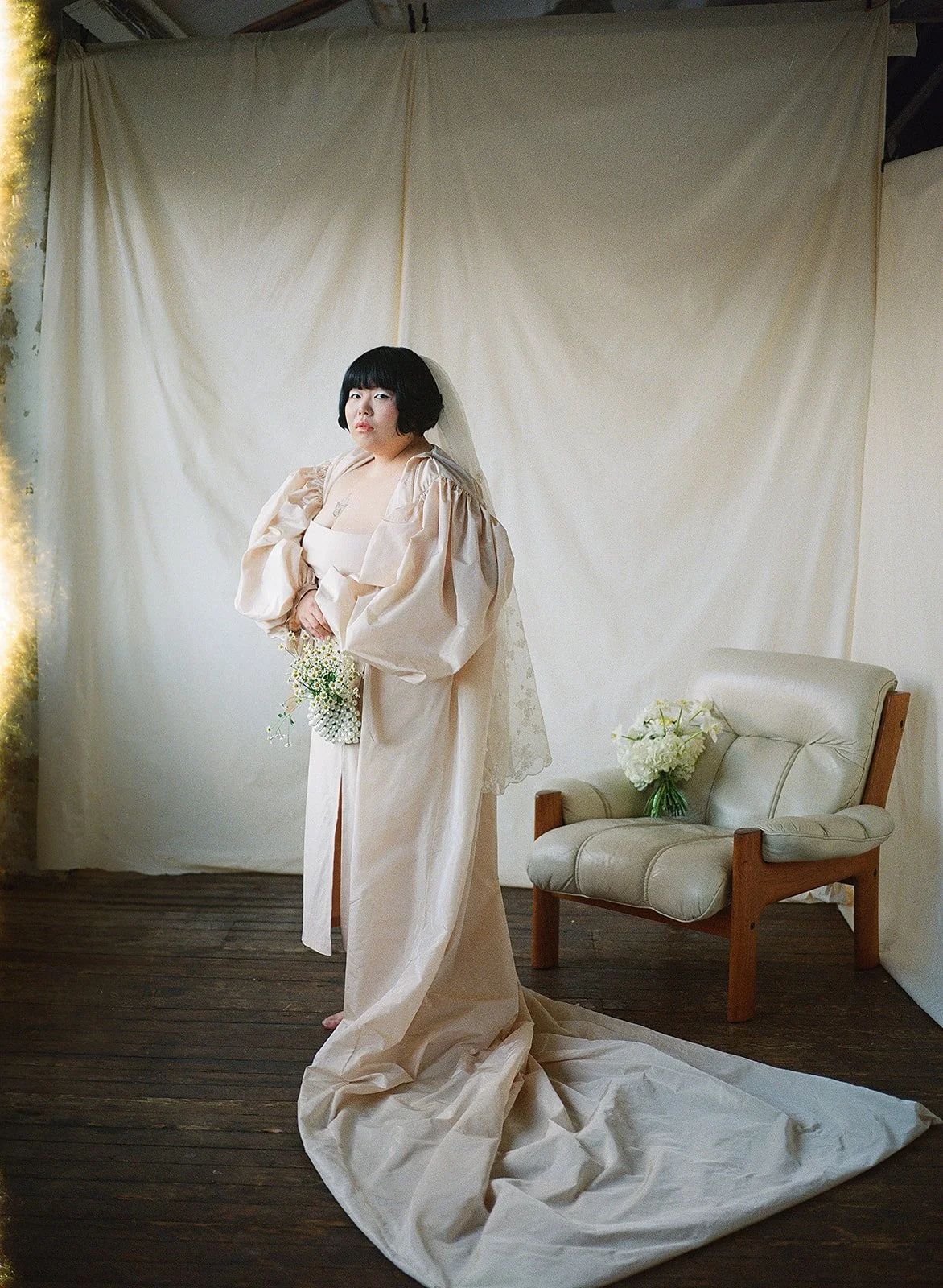 A person stands indoors against a softly draped fabric backdrop, wearing a long, pale gown with voluminous sleeves and a trailing hem that pools on the wooden floor. They hold a small bouquet of delicate white flowers at waist height. A light veil fa