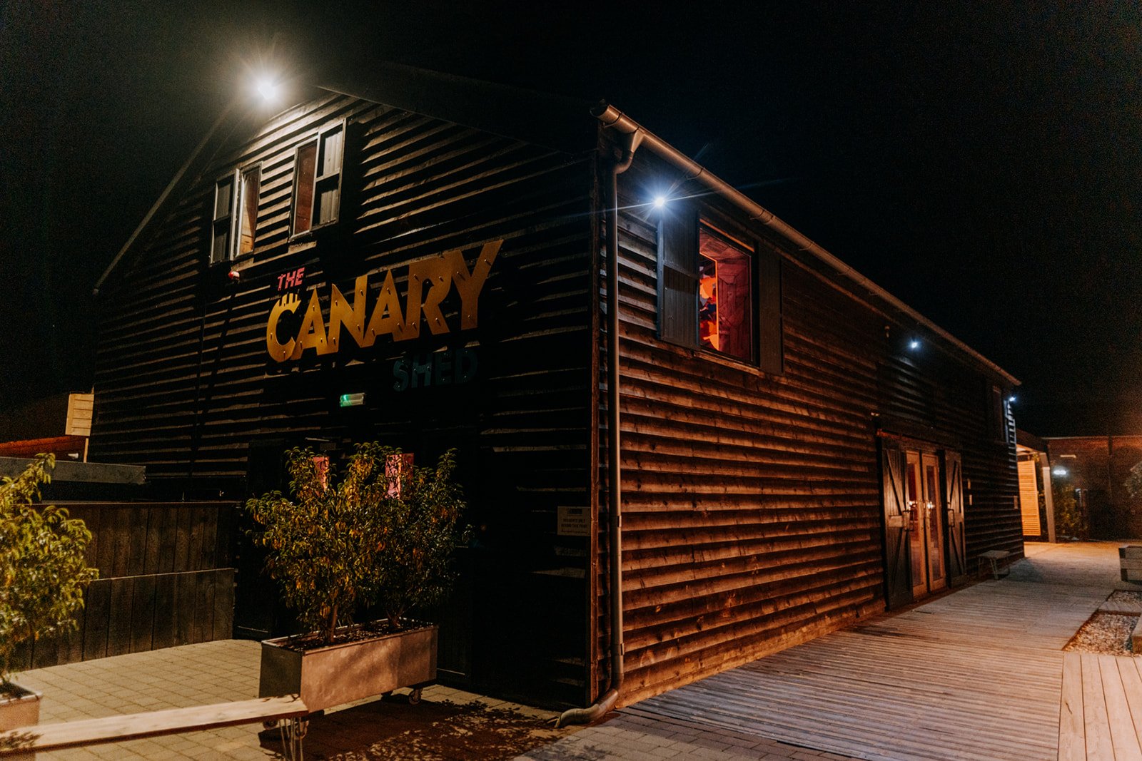 A dark timber clad building glows under night lights with The Canary Shed sign illuminated in bold yellow and blue lettering, warm light spilling from an upper window where colourful decorations are visible, and the surrounding walkway and planters s