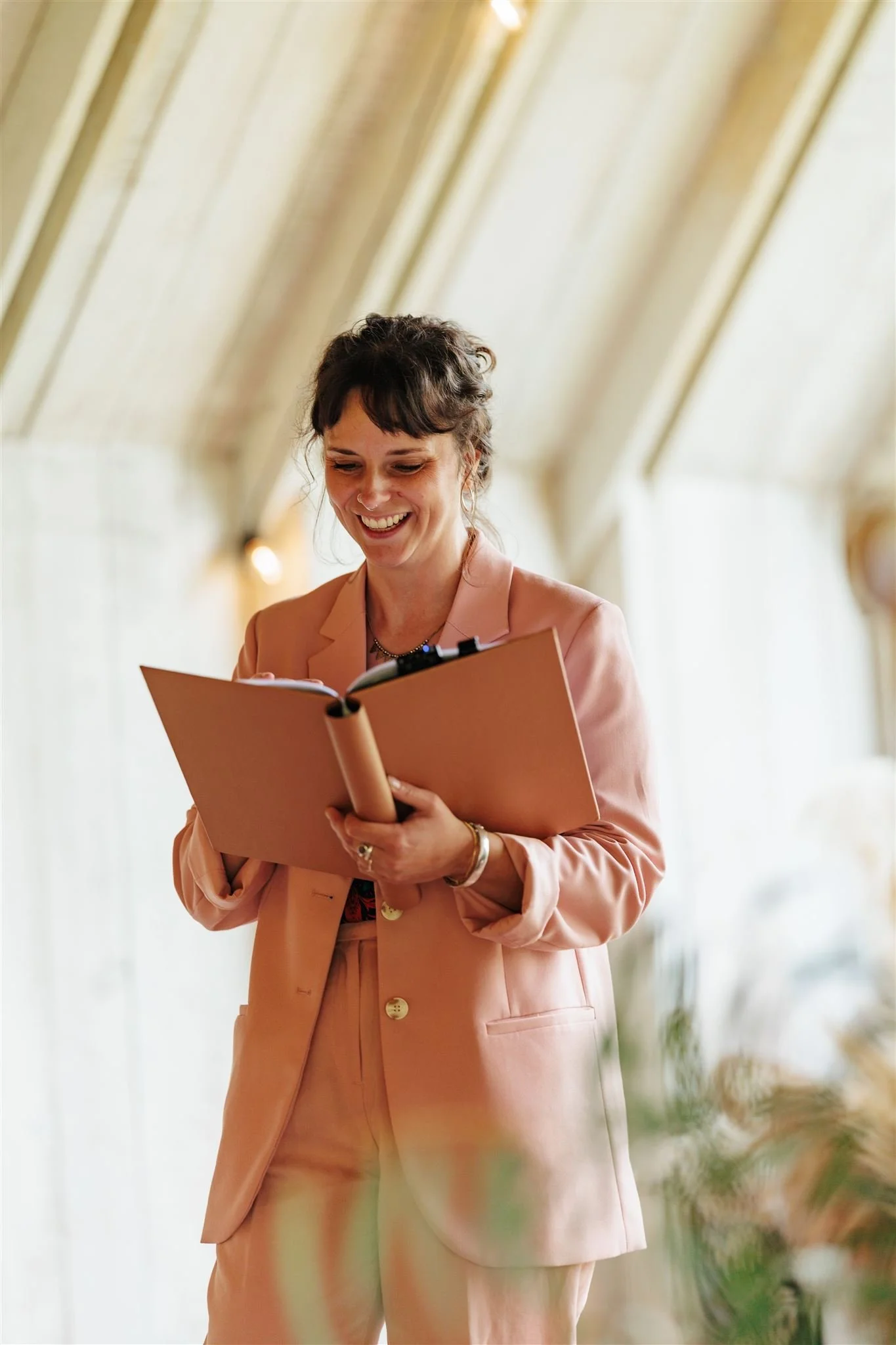 A smiling Cherry and Fern Celebrations in a blush pink suit reads from a ceremony book during an intimate indoor wedding. The soft natural light and elegant barn-style venue create a warm, romantic atmosphere. Ideal image for a UK wedding directory s
