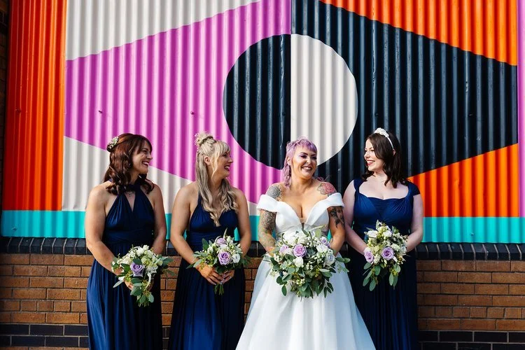 A colourful modern bridal party portrait captured by Rachel Burt Photography, featuring a bride with pastel purple hair and three bridesmaids in matching navy dresses standing against a bold geometric mural. The bright pink, orange, teal, and black s