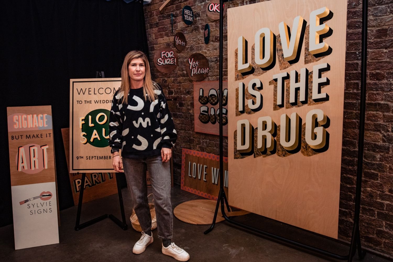 Hand-painted wedding signage by Sylvie Signs displayed at a wedding fair, featuring bold wooden signs including “Love Is The Drug” and welcome boards, shown with Esther the designer wearing a black patterned jumper, grey jeans and white trainers in a