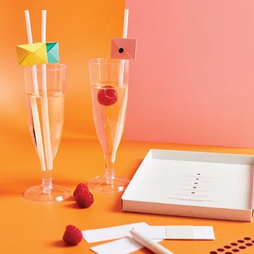 Two champagne glasses with origami-style drink toppers and raspberries, alongside a white Melon Teaspoon packaging box on a vibrant orange and pink background.