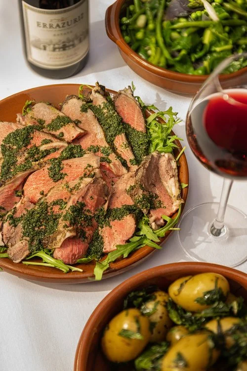 A beautifully presented wedding meal featuring sliced roast lamb topped with fresh herb pesto, accompanied by buttery new potatoes and vibrant green vegetables. A glass of red wine and bottle complete the elegant table setting, showcasing Tasting Pla