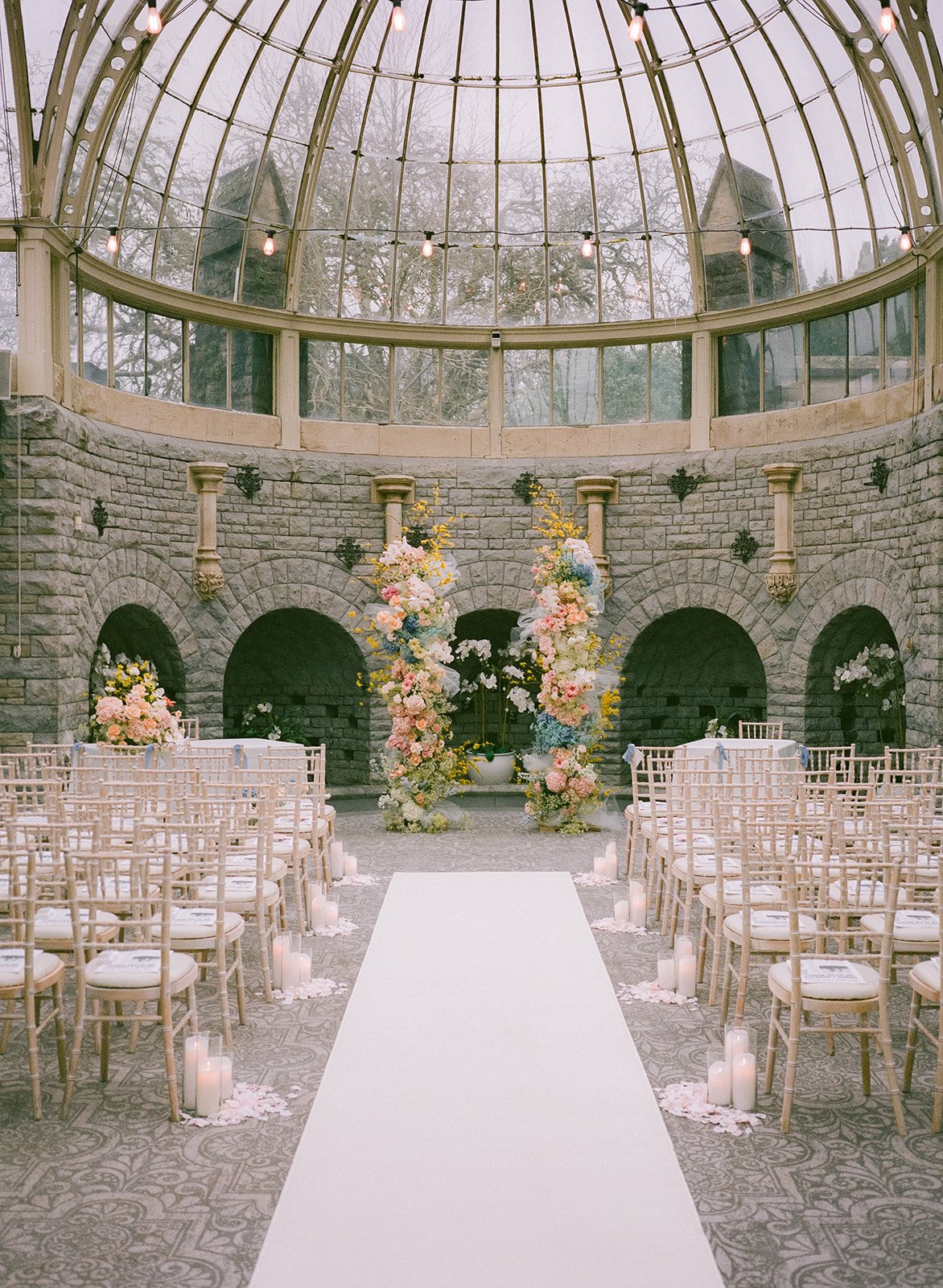 Elegant wedding ceremony setup by AMW Events in a grand glass-domed venue with pastel floral arrangements, white aisle runner, and candlelit decor. Xylle Frances Film