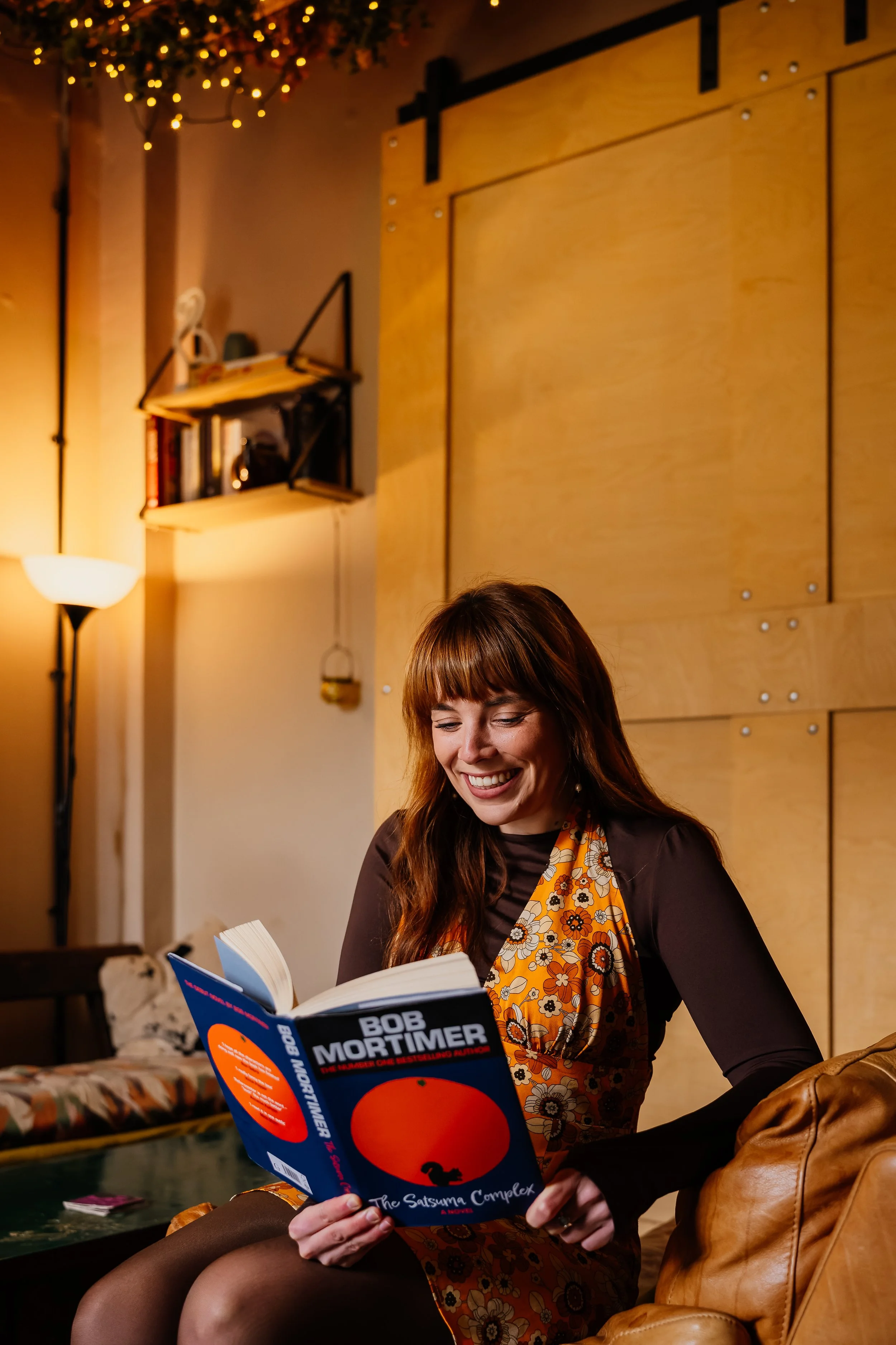 A smiling woman with red hair is reading a book by Bob Mortimer.