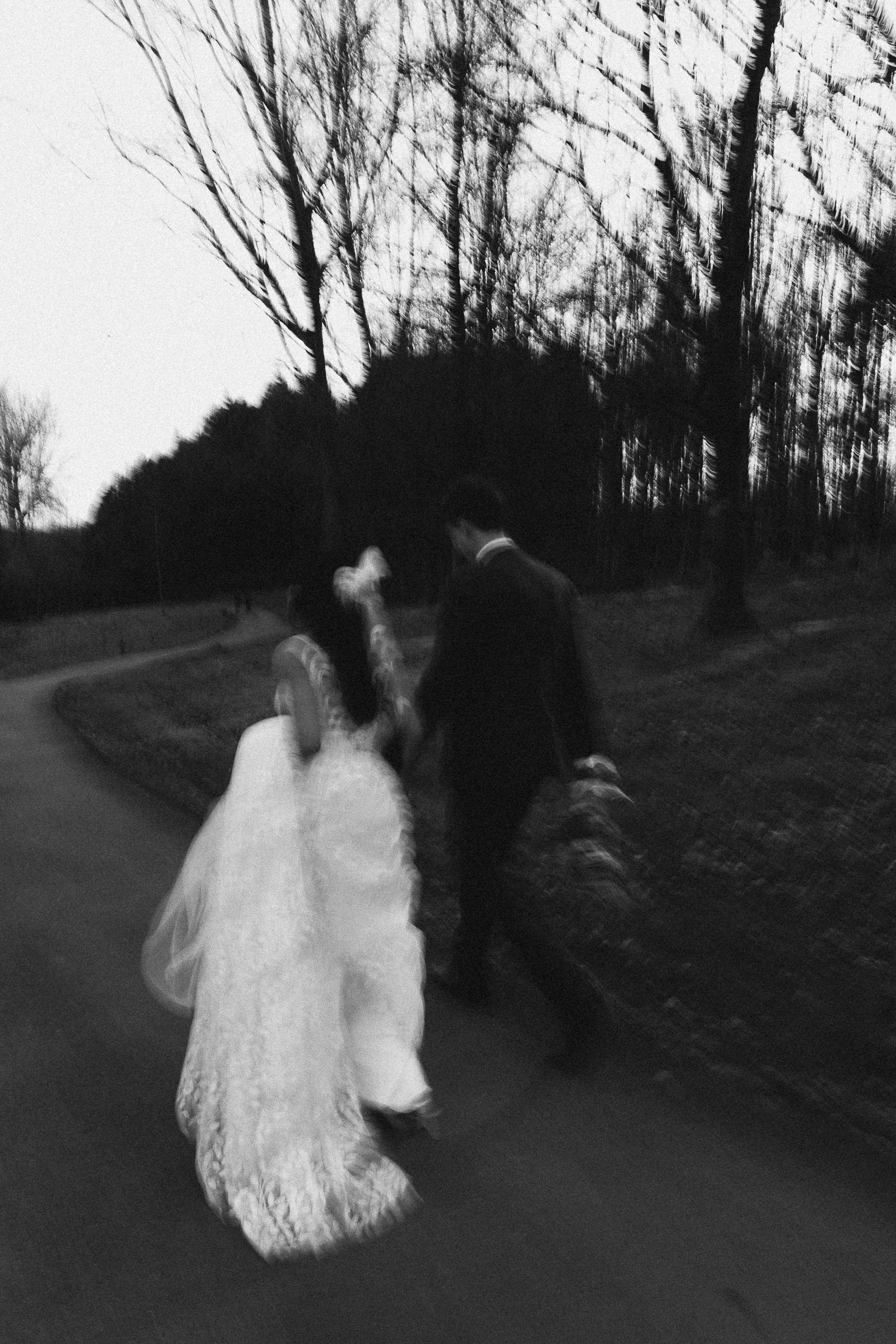 A beautifully artistic motion blur moment captured by YU Photography, showing the couple walking hand in hand along a winding path at dusk. The bride’s lace gown and flowing veil trail behind her as the groom keeps close beside her, both moving with 