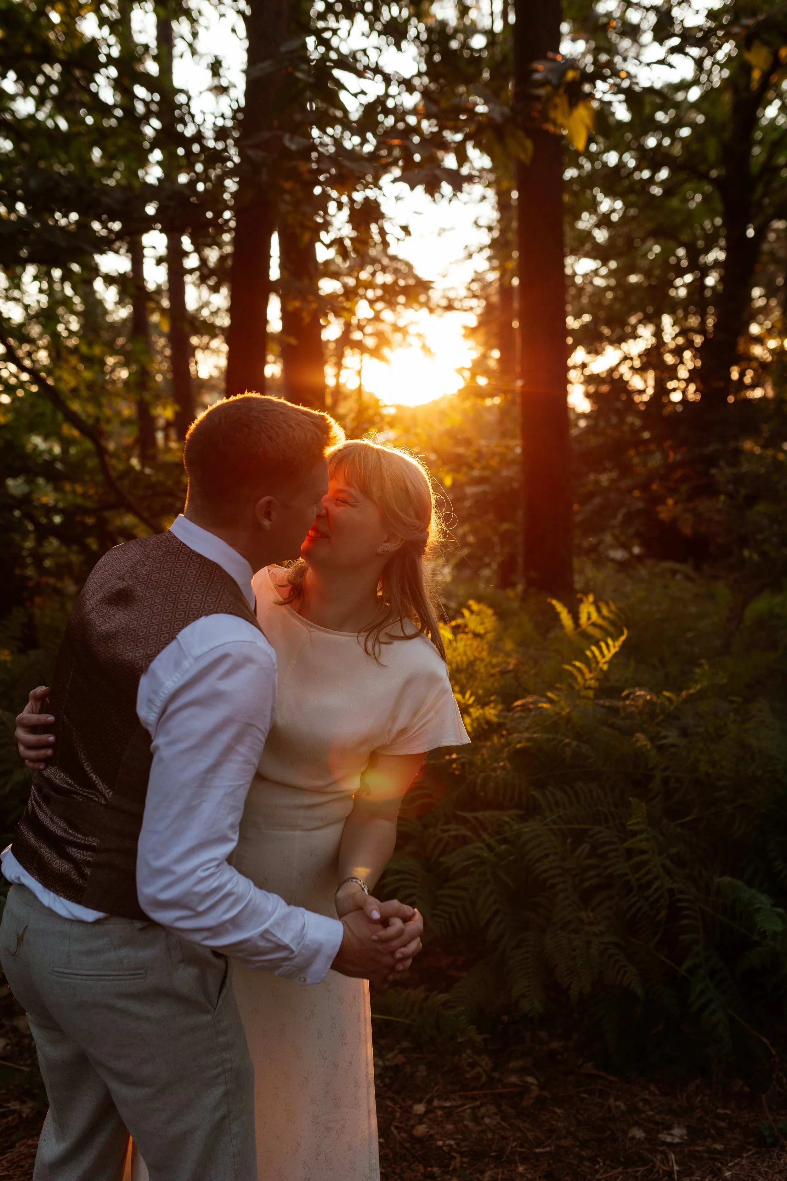 A warm, romantic wedding portrait captured by Laura Wilson Photography, showing a couple sharing an intimate moment in a sun-drenched woodland. The golden hour light filters through the trees, creating a soft glow around them as they hold each other 