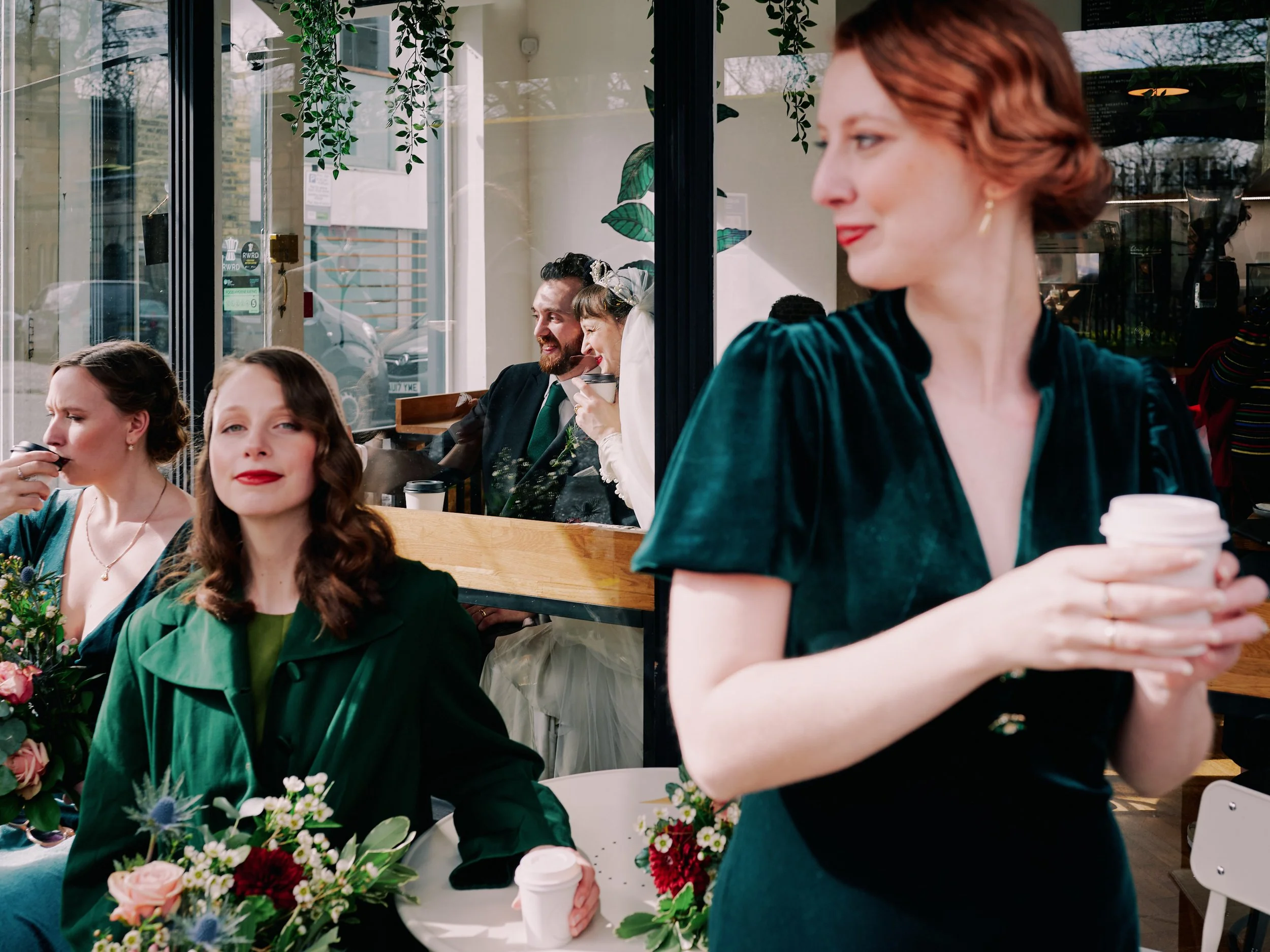 A lively and cinematic wedding moment captured by Wayne La Photography, showing the bridal party enjoying a calm pause outside a cosy café. Soft sunlight filters through the large windows, creating a mix of warm reflections and gentle shadows that gi