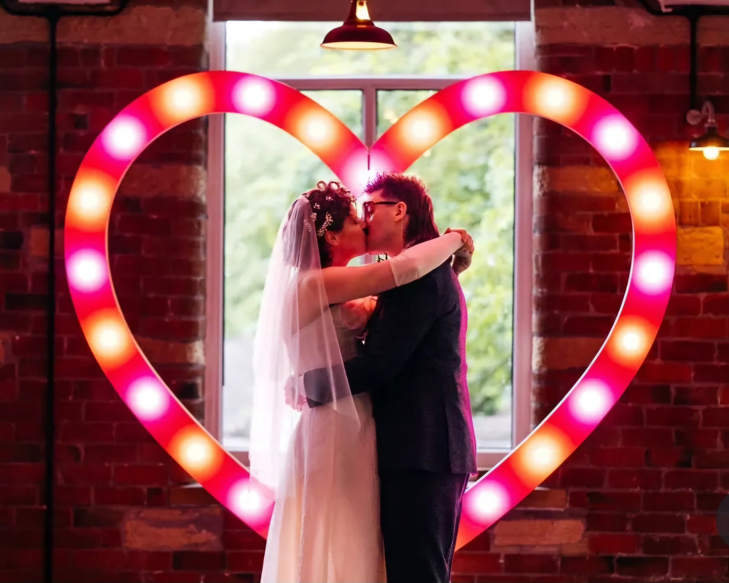 Couple kissing beneath a large illuminated heart prop, creating a bold and playful wedding photo moment. Statement heart light hire ideal for alternative weddings, fun reception styling and unforgettable photo backdrops.