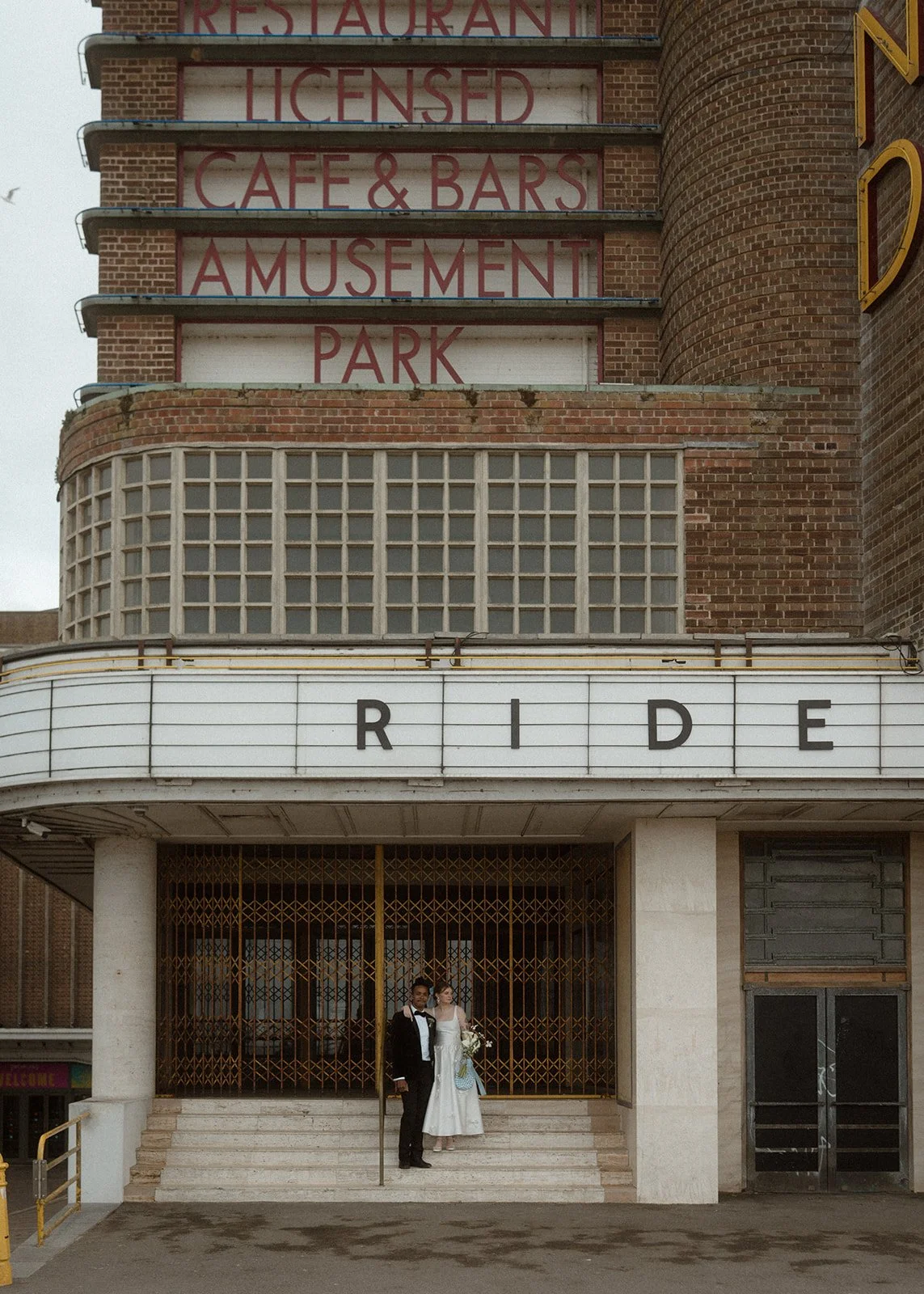 A couple stands at the base of an old seaside amusement building with bold retro lettering and weathered brick textures while the marquee above them spells out RIDE and the closed golden gates behind them frame their formal attire which consists of a