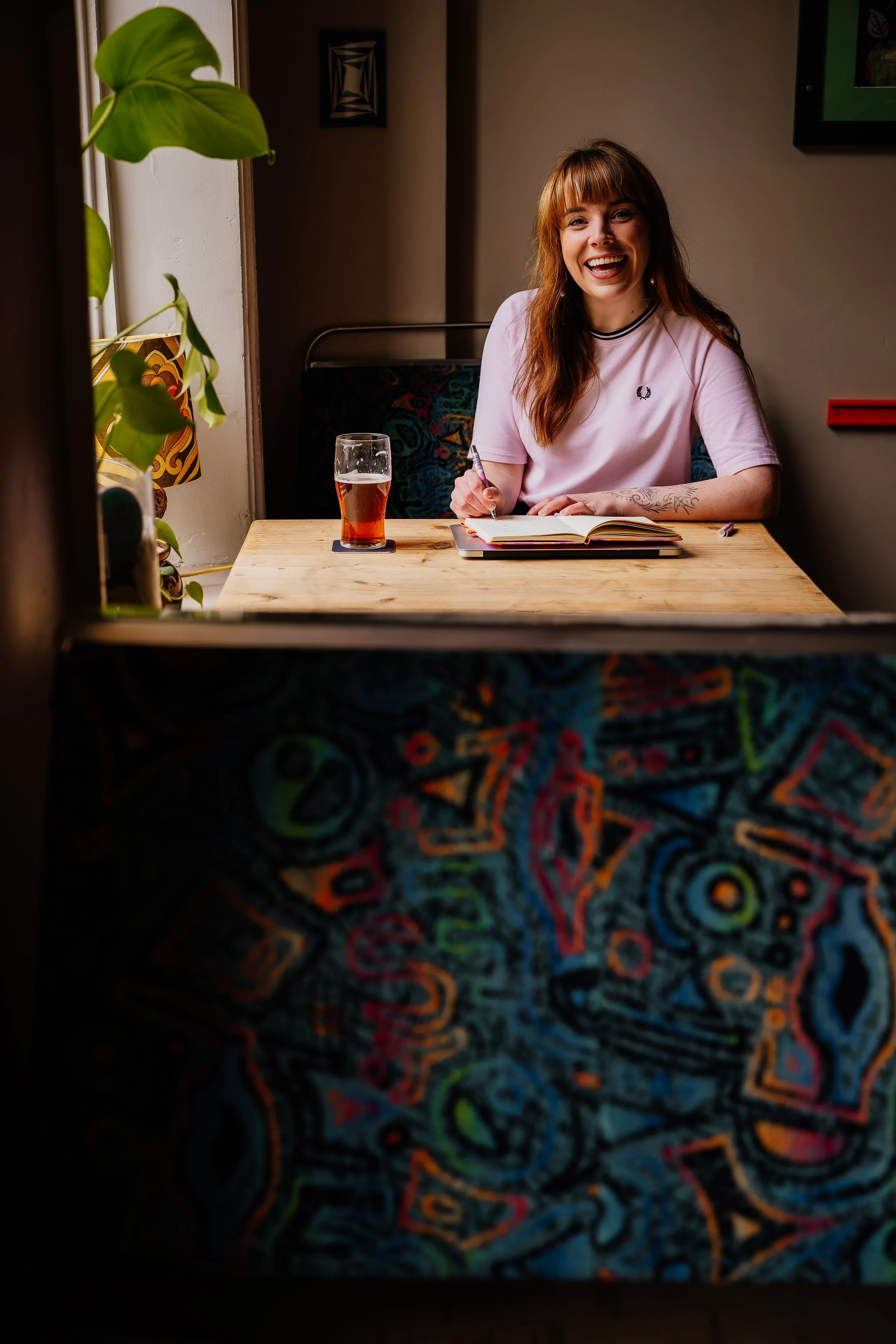 Abi aka Full Of Soul Celebrant is sitting by a window, writing some bespoke wedding vows, with a pint of beer by her side.