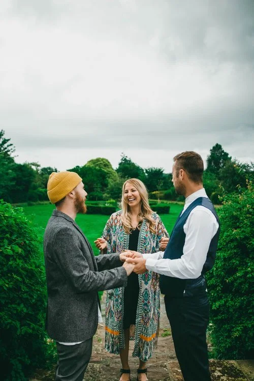 A joyful outdoor wedding ceremony led by Roxy Celebrates Love, as two grooms exchange vows surrounded by lush green countryside. The relaxed, modern ceremony captures the warmth and inclusivity of contemporary UK weddings, with the celebrant dressed 