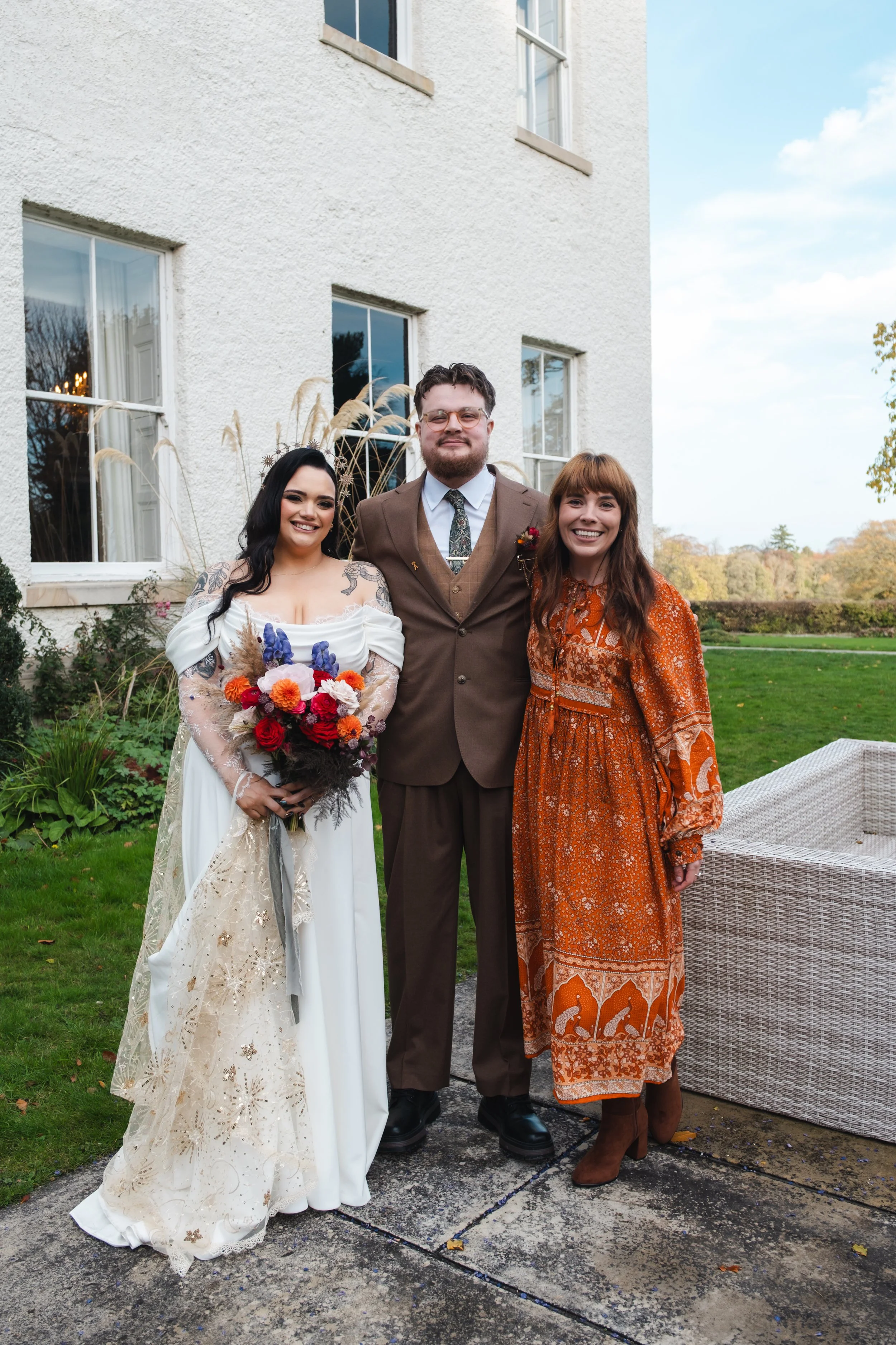 A celebrant with a beautiful, floral dress and long, red hair, is standing next to her bride and groom.