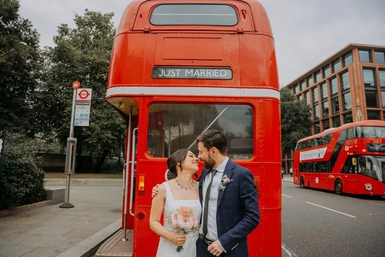 A newly married couple share a kiss in front of a classic red London bus displaying a Just Married sign, captured by Joshua Humphrey Photography. Their joyful moment and the iconic city backdrop create a vibrant urban wedding portrait full of charact