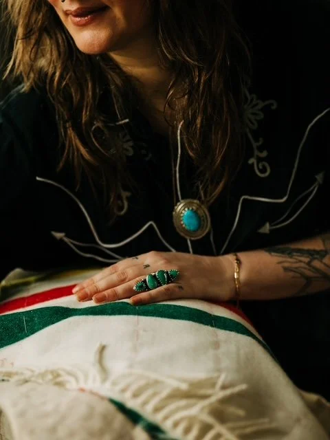 A close-up view shows Kellye from Long Time Gone wearing a black Western-style shirt with white embroidered detailing, along with a large turquoise pendant and a distinctive multi-stone green ring. Her hand rests on a cream blanket with red, green an