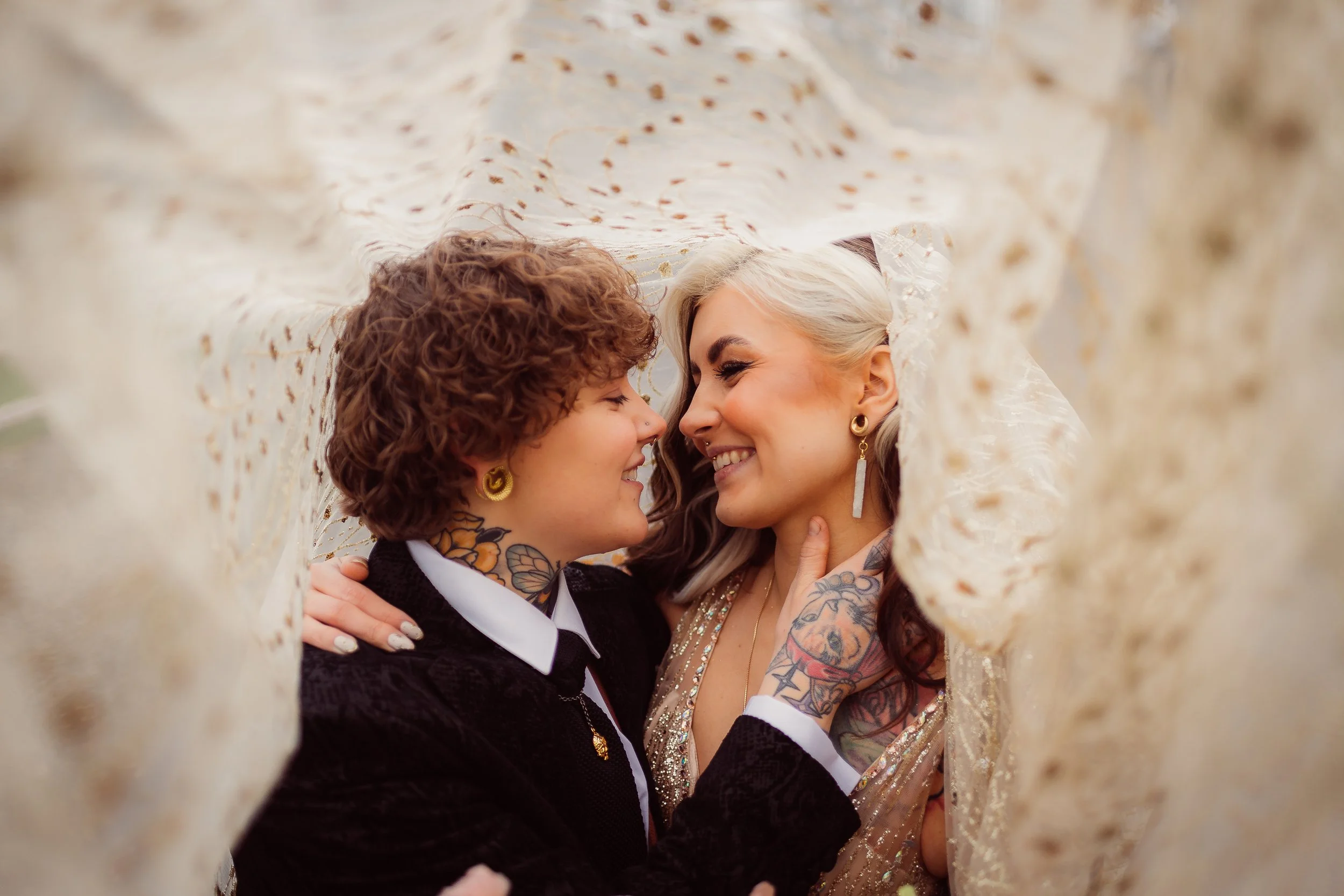 A couple share an intimate joyful moment wrapped beneath a delicate embroidered veil, captured by Gemma Gaskins Photography. Their smiles, tattoos and stylish outfits create a warm modern wedding portrait full of personality, romance and creative art
