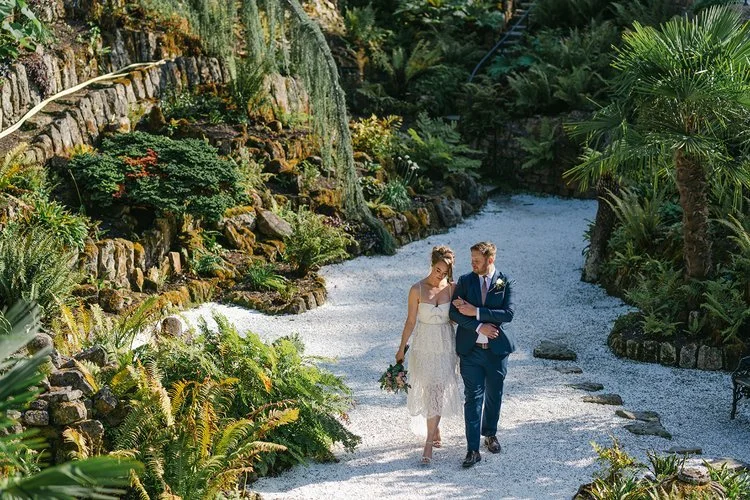 A bride and groom walk arm in arm along a sunlit garden path surrounded by vibrant ferns, moss-covered rockeries and tropical greenery, beautifully captured by Christy Photography. The peaceful setting, natural textures and warm light create a romant