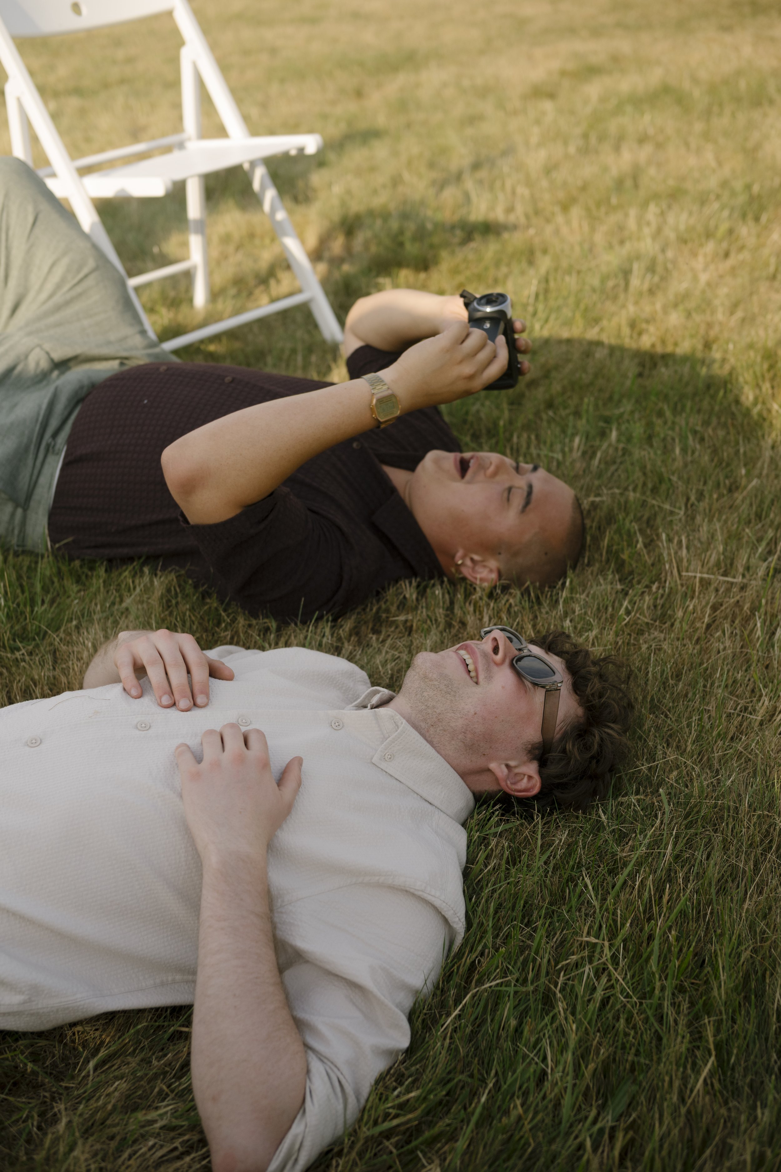 A relaxed and candid wedding moment captured by Roz Pike Photography shows two guests lying back on sunlit grass during a summer celebration, one holding a camera while laughing as the other rests beside them wearing sunglasses, perfectly reflecting 