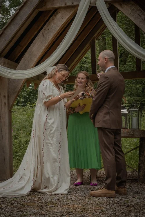 Outdoor wedding ceremony under a rustic wooden arch draped with white fabric. The bride, wearing an embroidered cream gown, laughs as she exchanges vows with the groom in a brown suit, while the celebrant, dressed in a green skirt and floral blouse, 