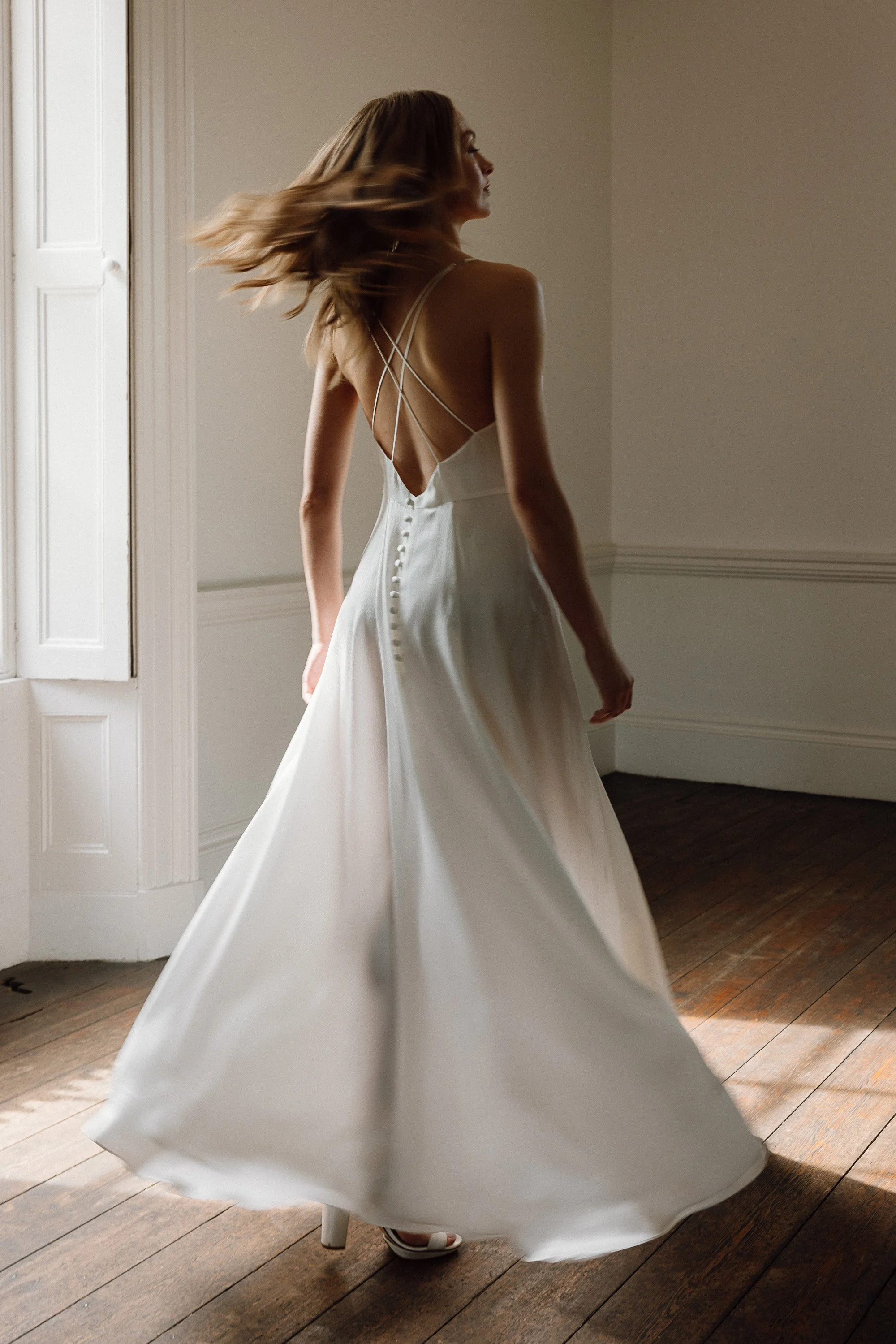 Soft daylight pours through the tall window as the bride twirls across the wooden floor, her silk gown sweeping in a wide circle and revealing a low back framed by delicate crisscross straps and a row of covered buttons that trail gracefully down the